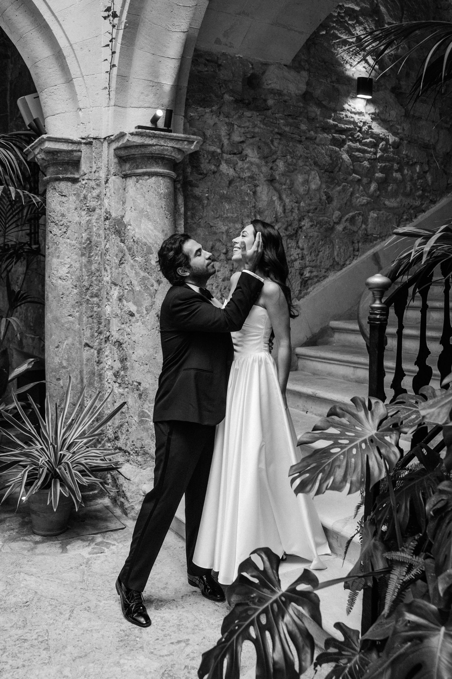 A couple in formal attire dances playfully in a stone courtyard with tropical plants and arched architecture.