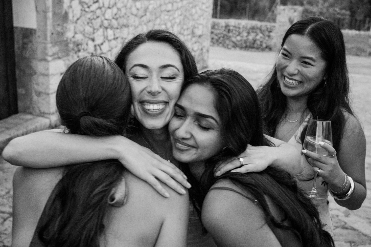 Black and white photo of friends sharing an emotional embrace while another holds a wine glass in the background.