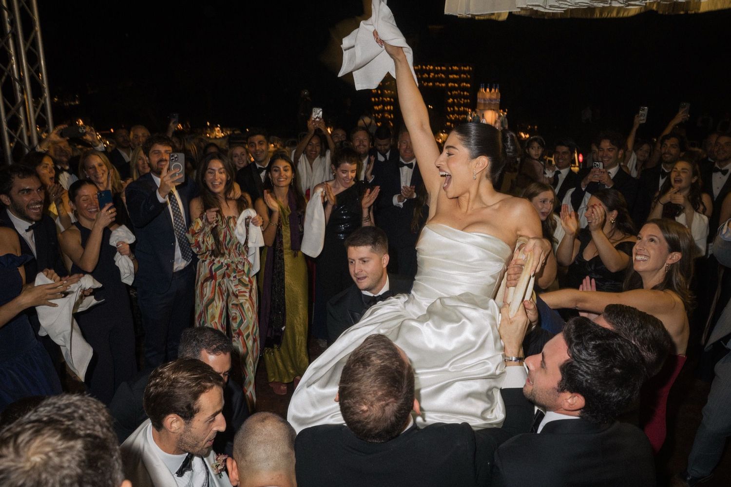 Wedding celebration at night with a bride in white satin dress being lifted up by guests on a crowded dance floor.