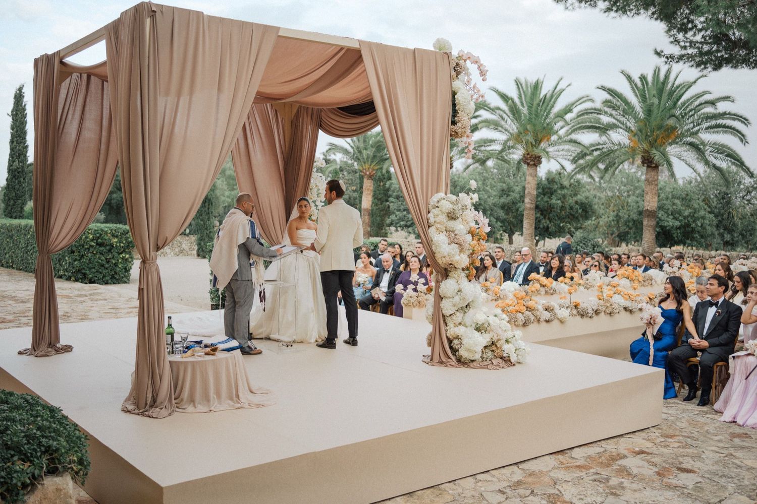 Elegant outdoor wedding ceremony under a beige draped canopy with palm trees and floral decorations in the background.