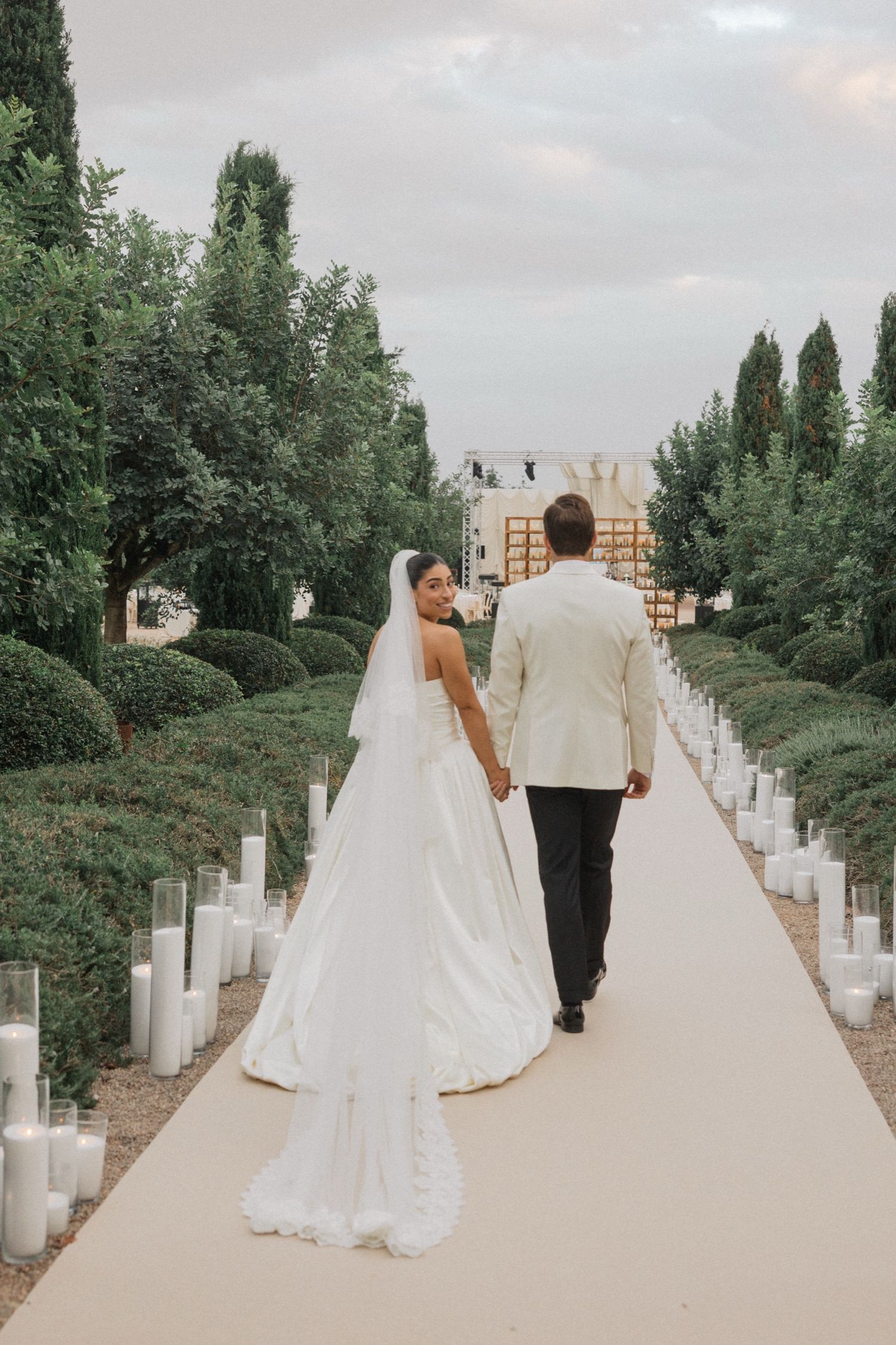 A bride and groom walk hand in hand down a white path surrounded by green trees and cemetery headstones.
