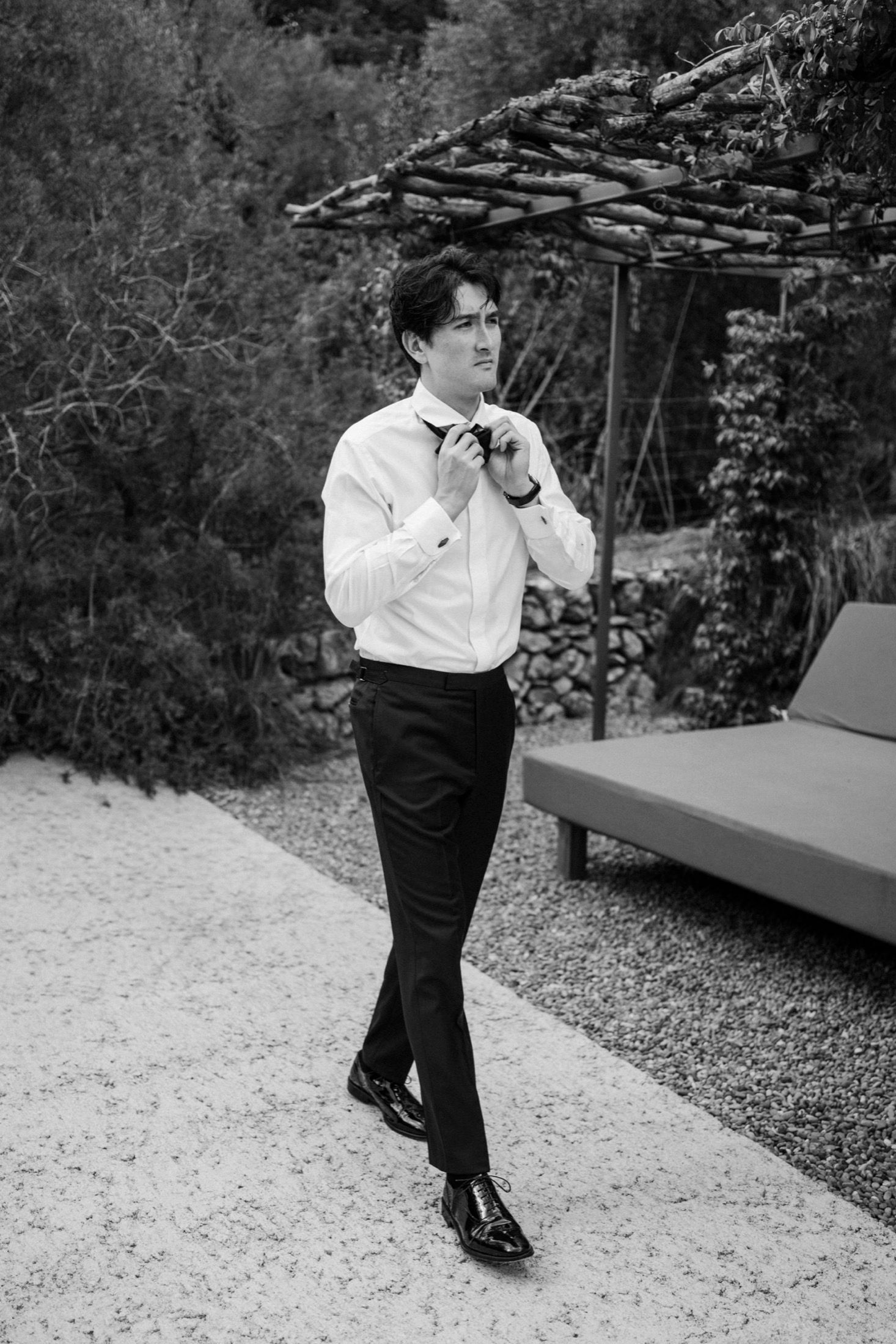 Black and white photo of someone in formal attire walking outdoors near a garden pergola and lounge chair.