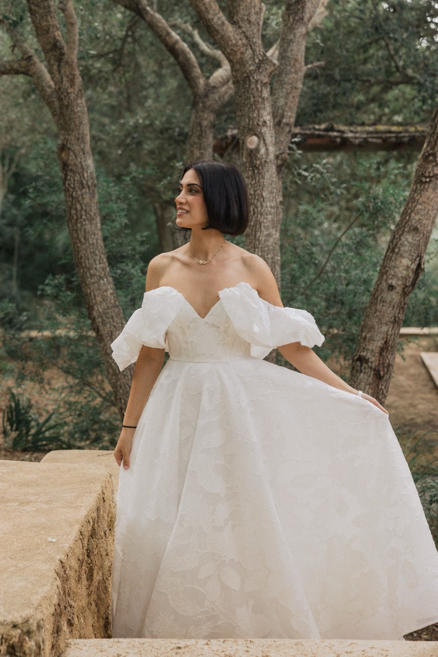 A bride in an elegant off-shoulder white gown with puff sleeves poses outdoors among trees in a natural setting.