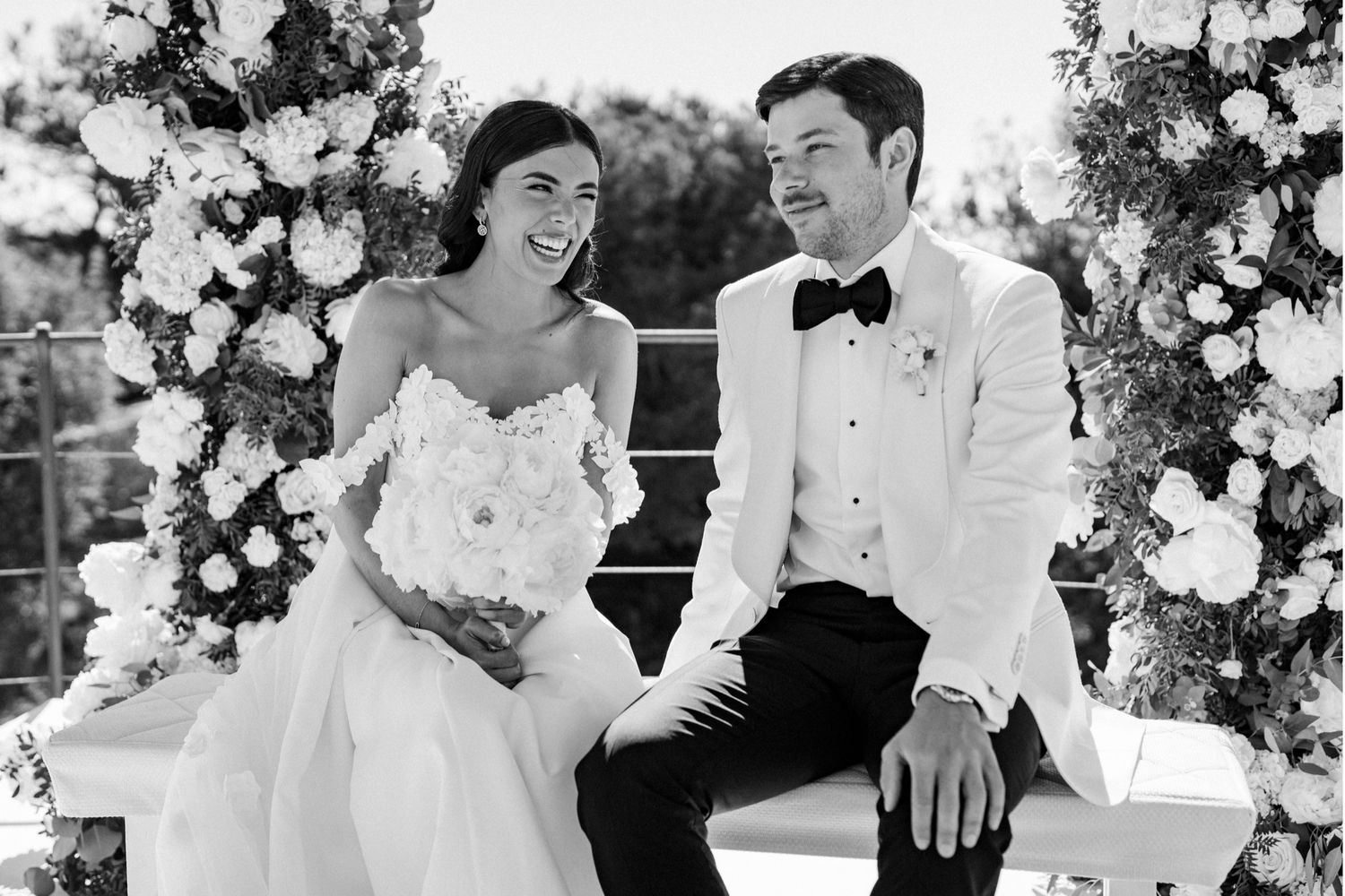 Black and white wedding photo of smiling bride in strapless gown and groom in white tuxedo seated among floral arrangements.