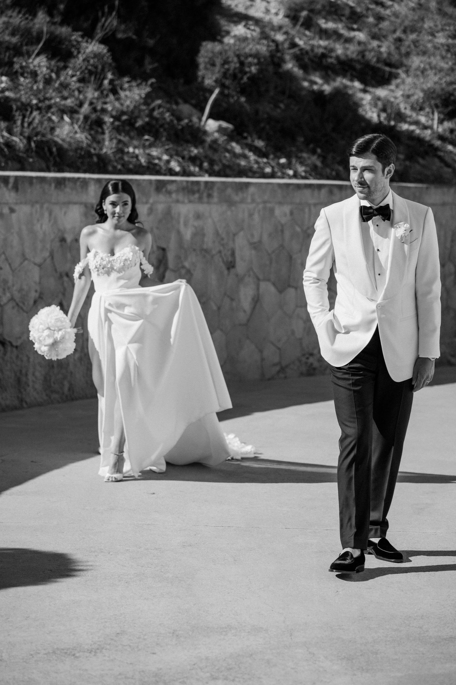 Black and white photo of bride and groom walking outdoors in elegant wedding attire.