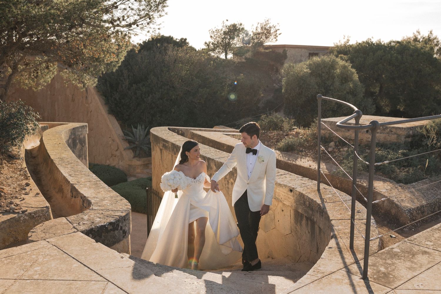 Bride and groom hold hands walking on stone steps at golden hour outdoor wedding venue.