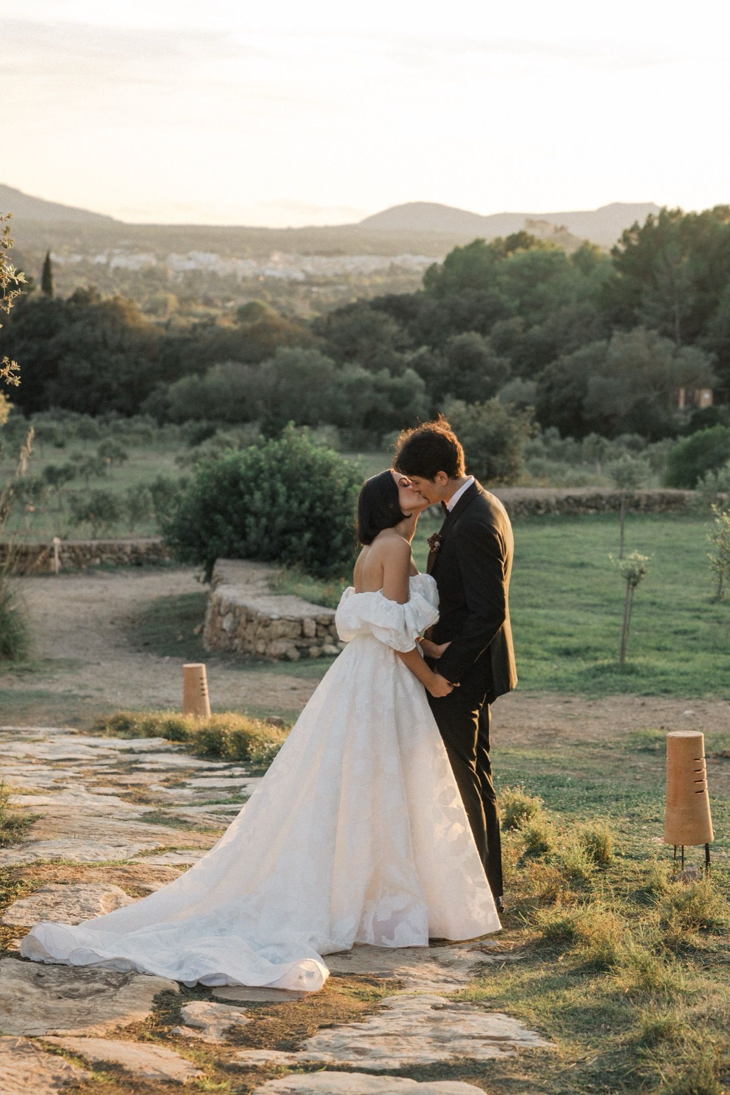 Newlyweds share an intimate moment at sunset in a scenic countryside setting with mountains in the background.