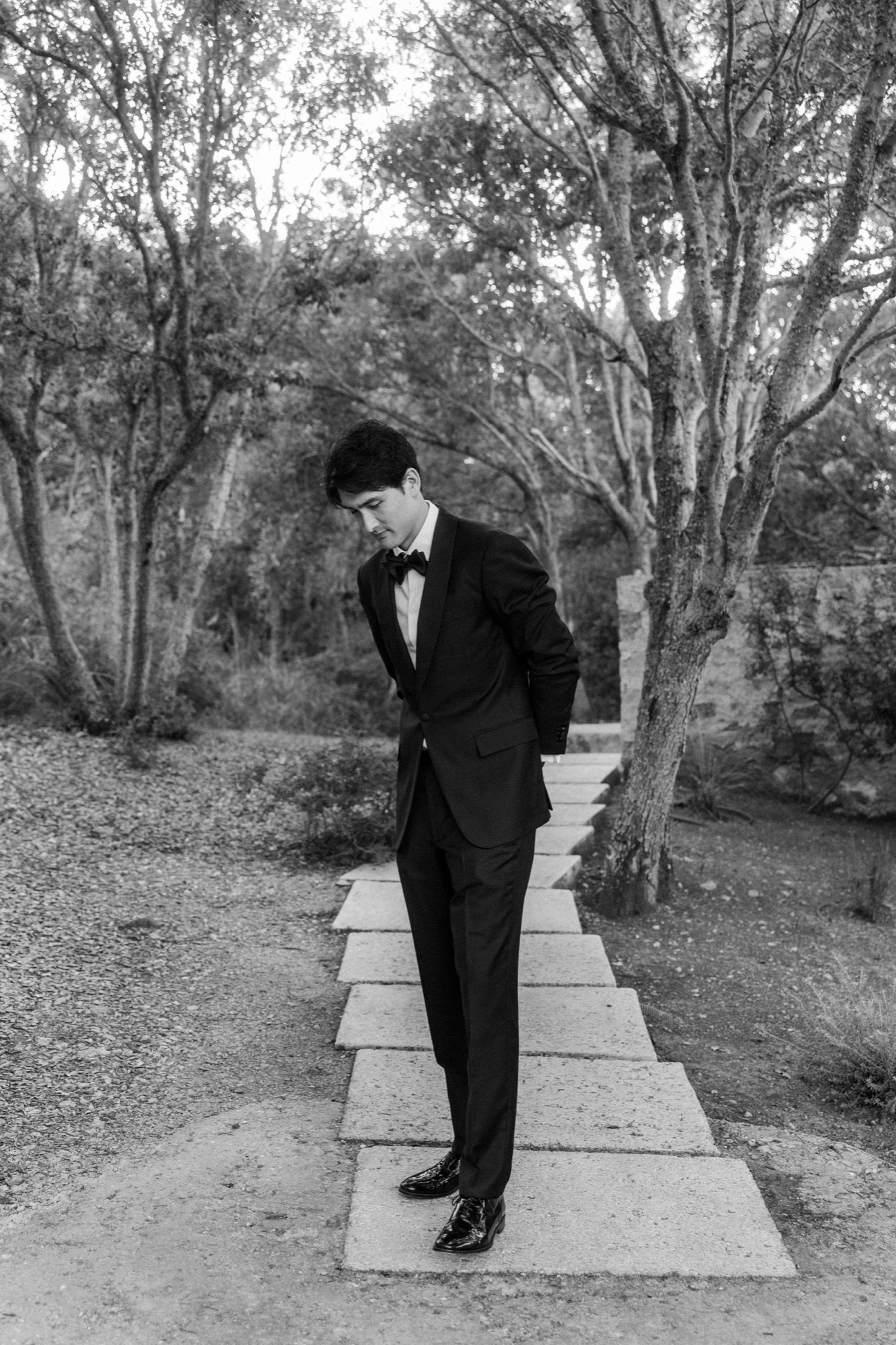 A figure in a black suit stands contemplatively on concrete steps surrounded by trees in a black and white photograph.