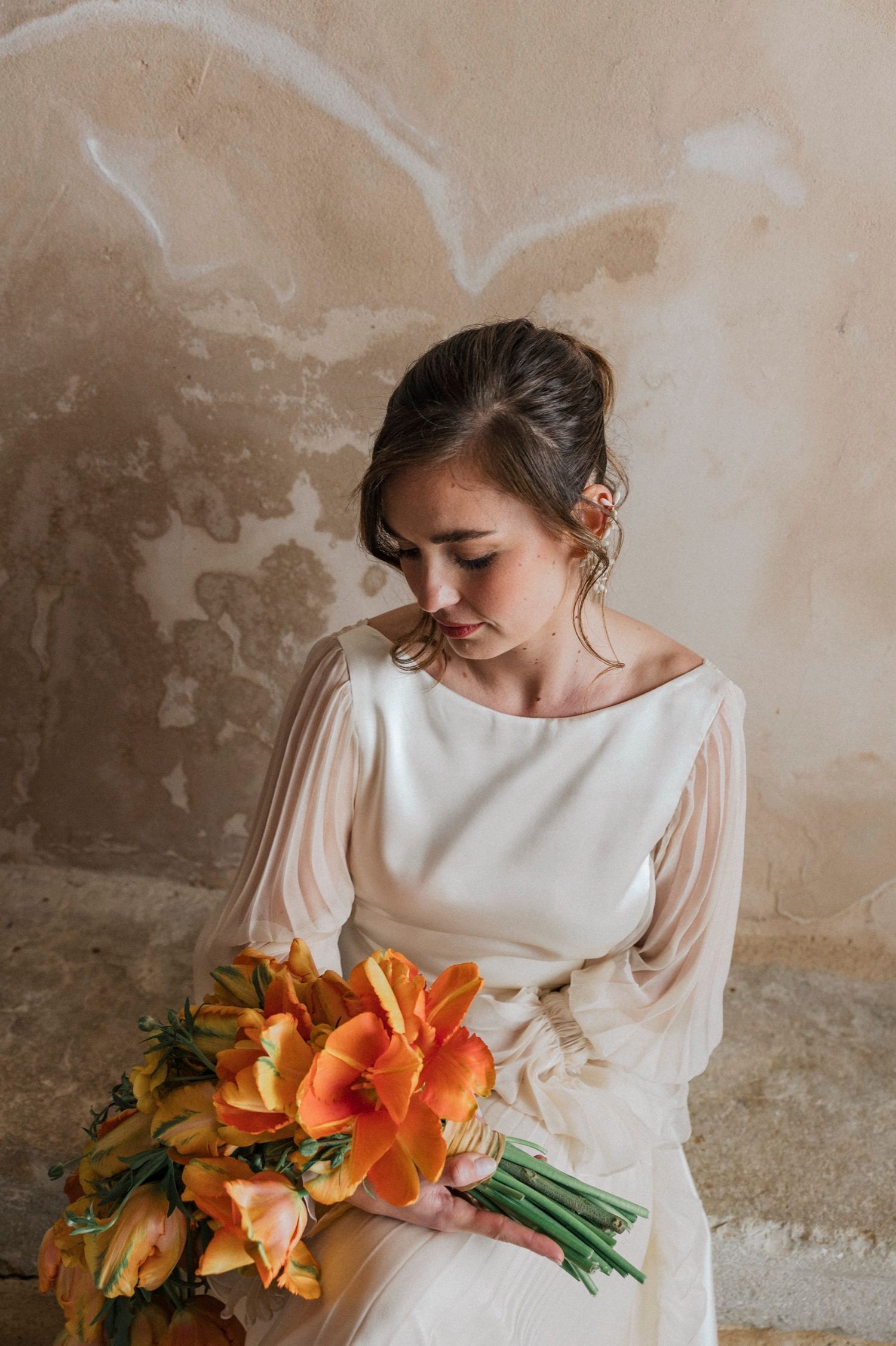 A bride in a classic white long-sleeved dress poses with vibrant orange lily bouquet against a textured wall.