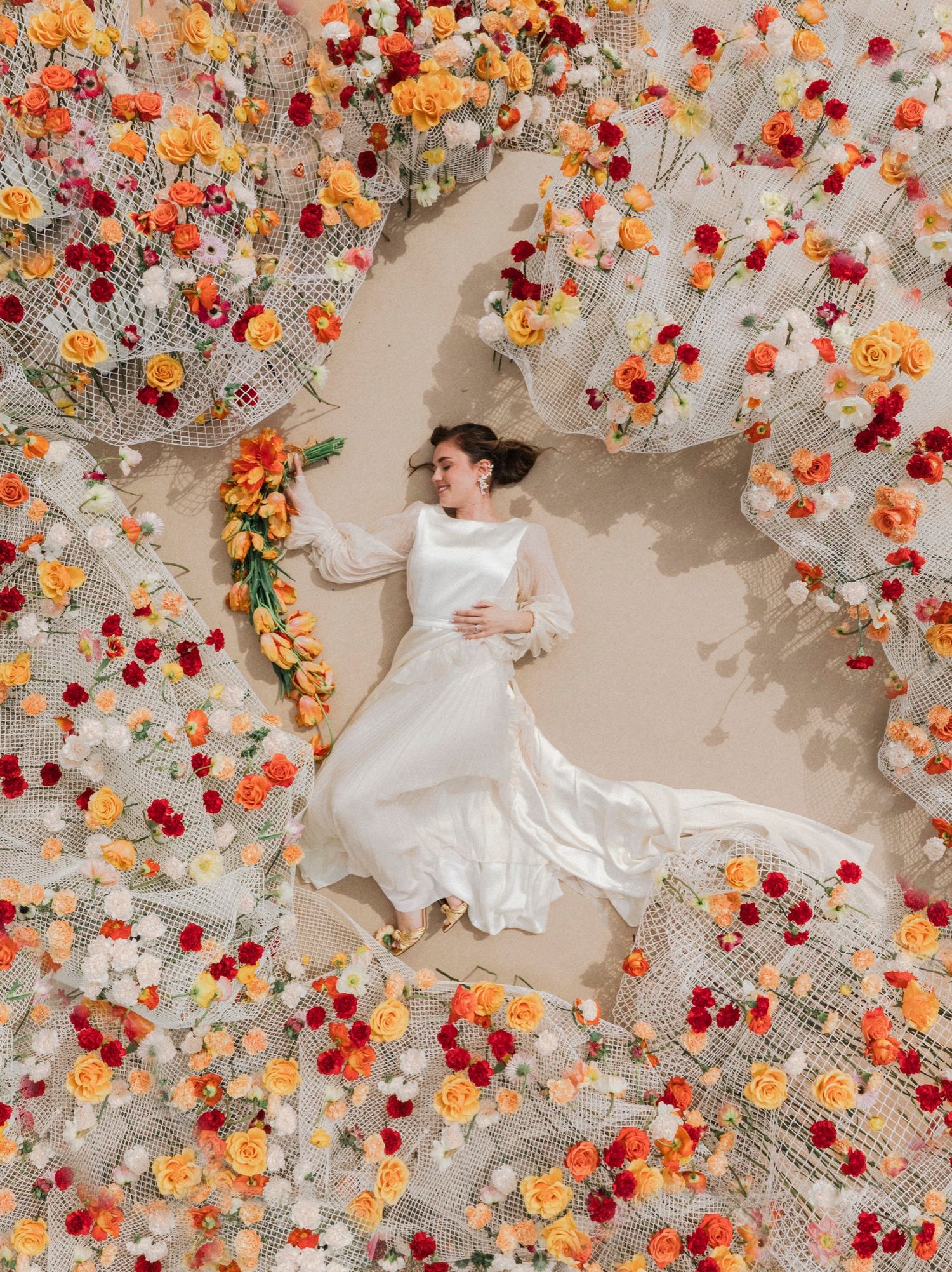 Artistic overhead view of a person in white dress lying on floor surrounded by scattered orange and red flowers.