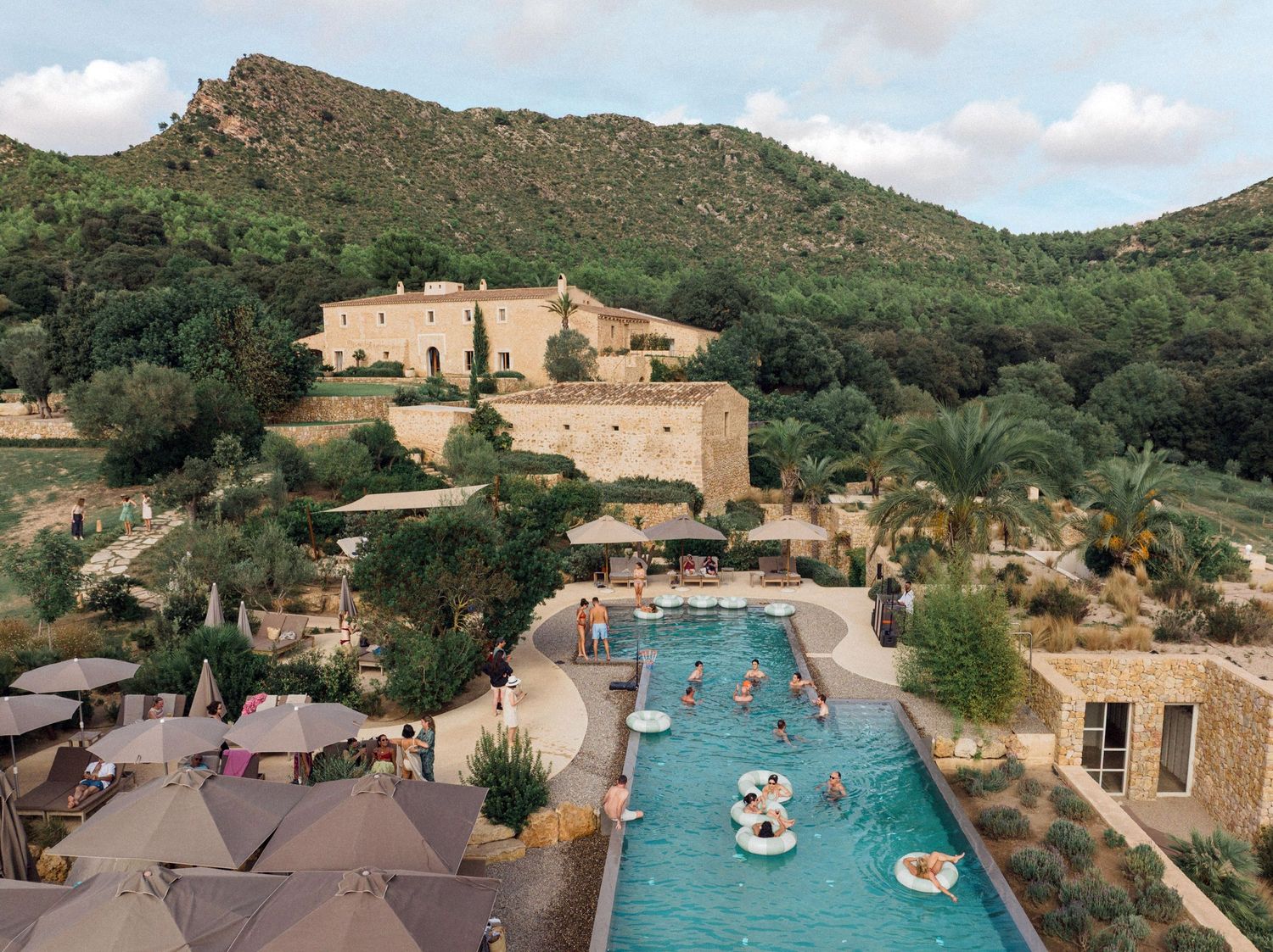 Aerial view of a luxury resort swimming pool nestled in the mountains with surrounding stone buildings and landscaping. Es Racó d'Artà