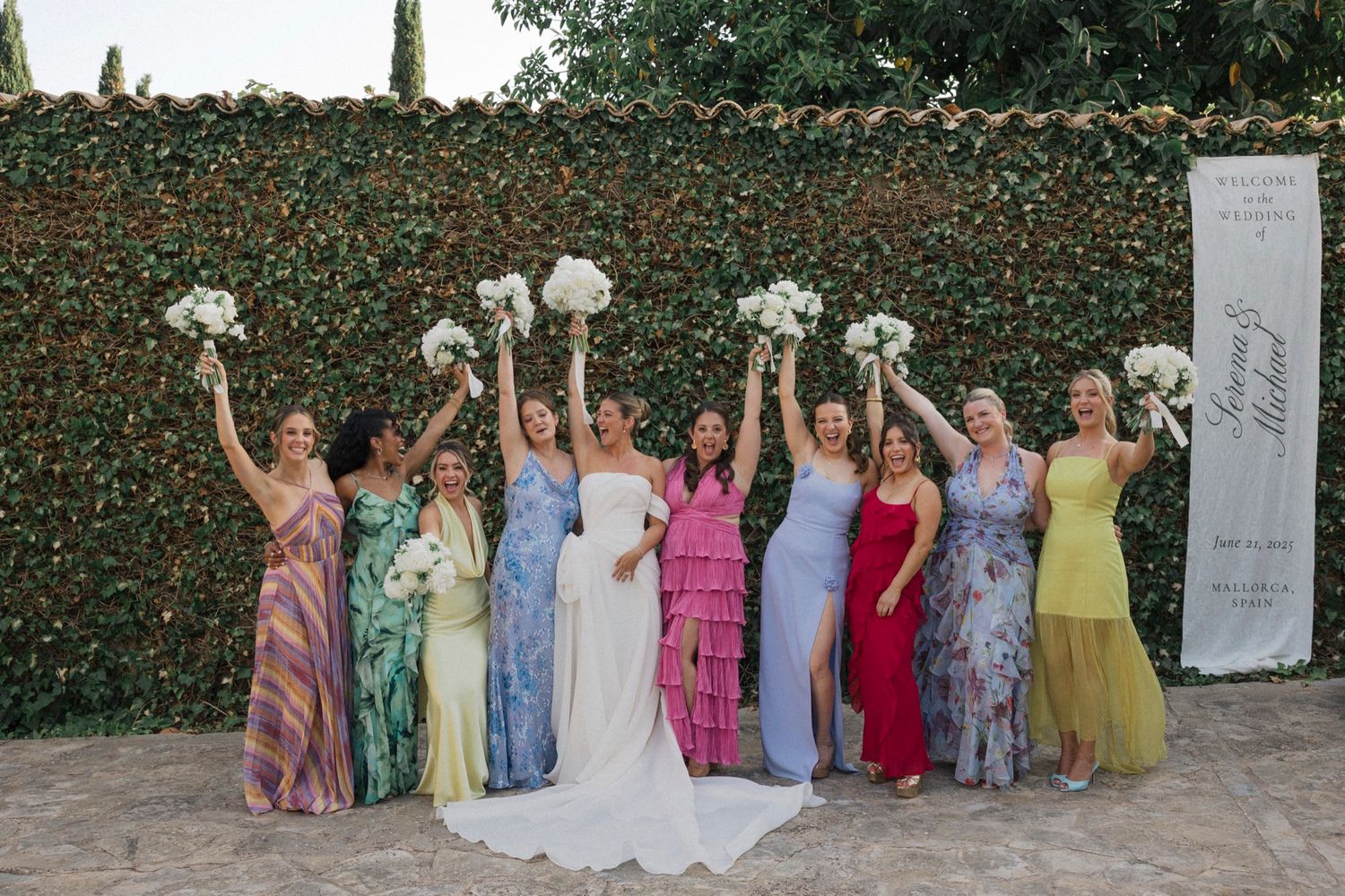 Bridesmaids in pastel dresses line up against garden wall for group photo.