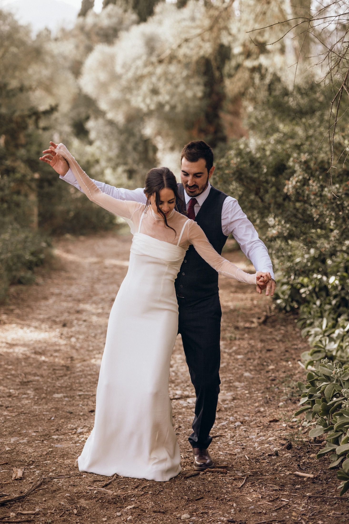 A couple in wedding attire dances playfully on a dirt path surrounded by lush greenery and trees.