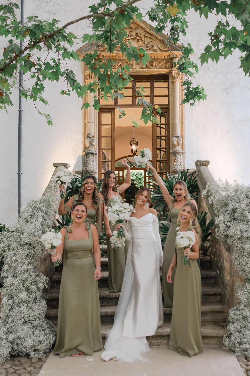 Wedding party in olive green dresses and white gown pose joyfully under green foliage.