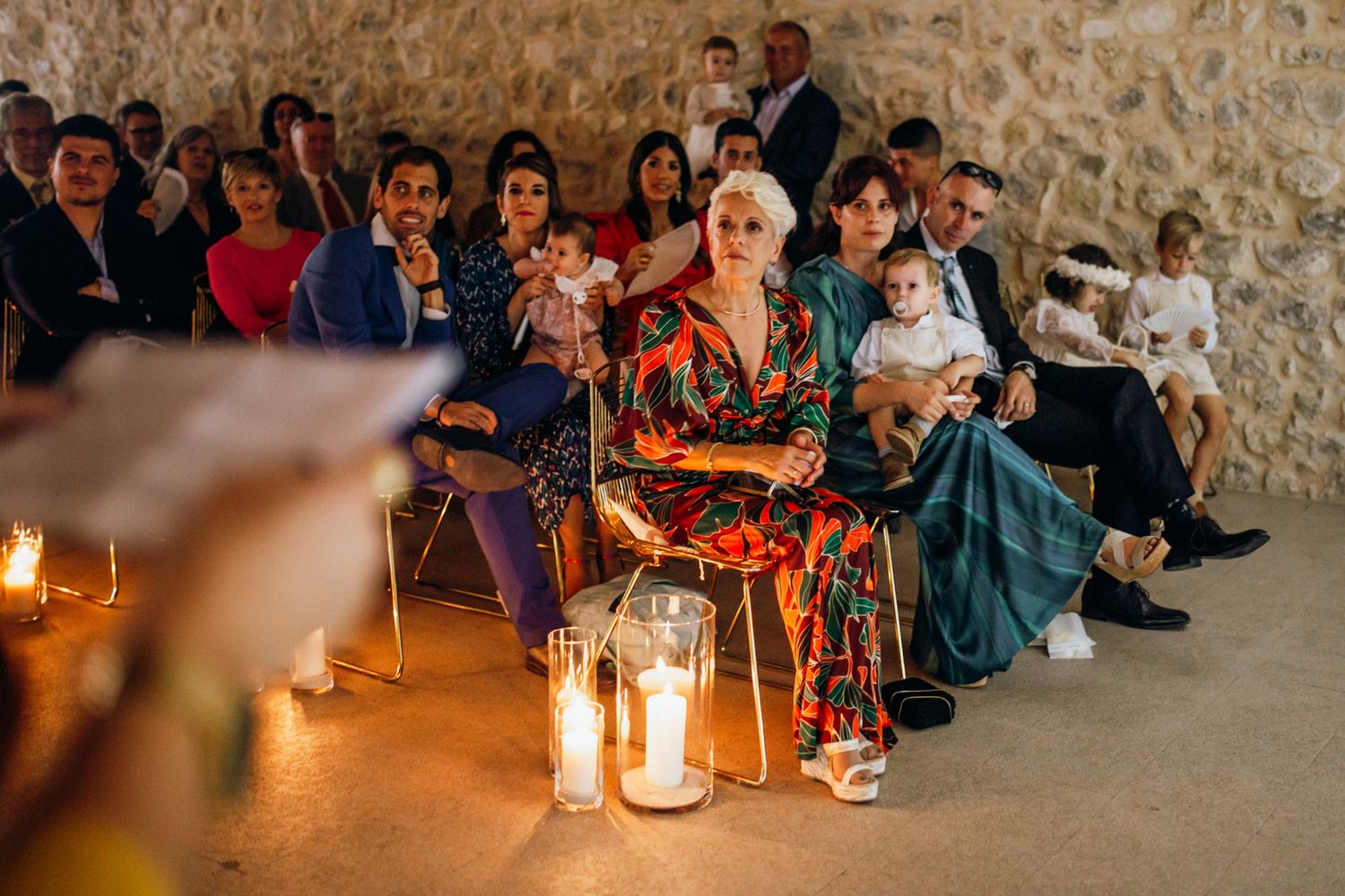 Wedding guests seated on stone benches with glowing lanterns creating warm ambiance at evening ceremony.