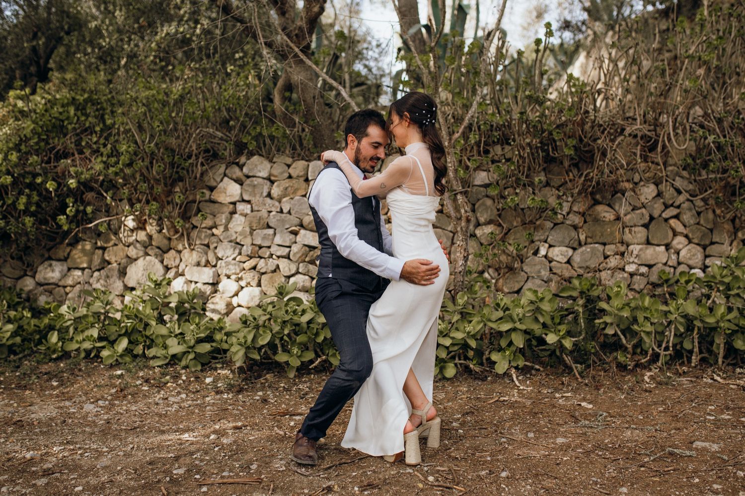 A romantic outdoor photo session featuring a couple in wedding attire embracing near stone walls and natural foliage.