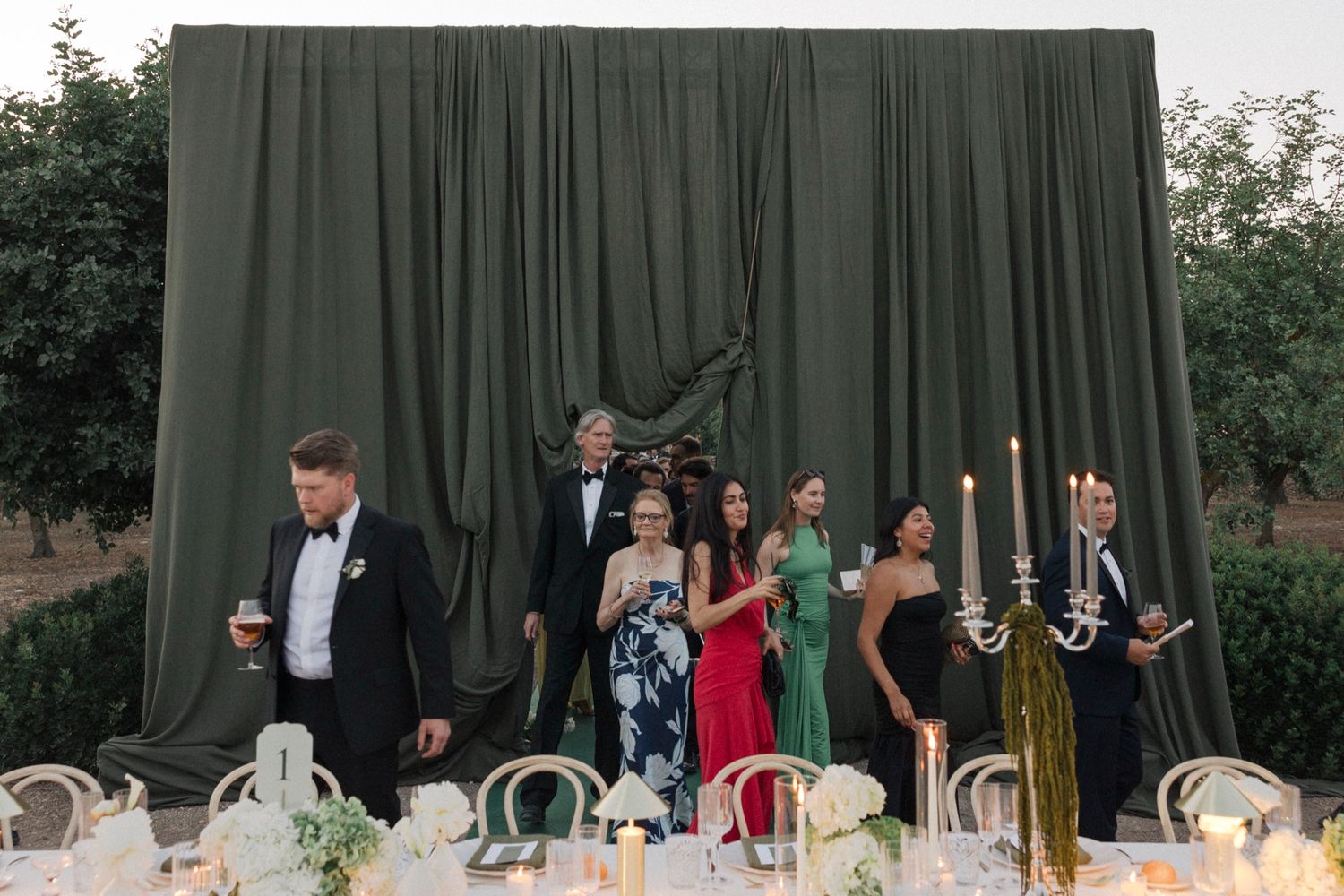 Wedding reception tables set against dark green curtain backdrop with candlelight.