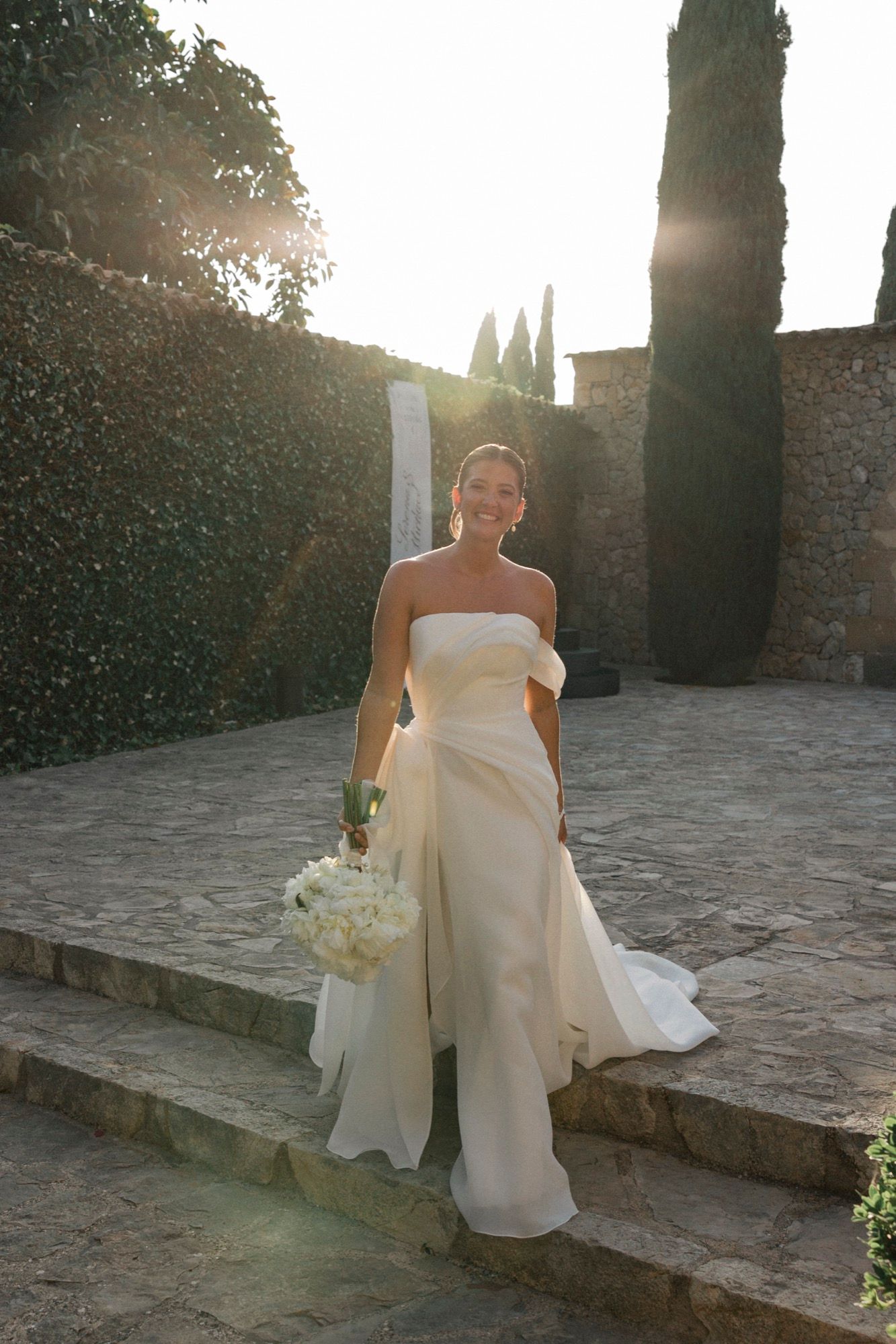 Bride in strapless white gown poses in sunlit stone courtyard with bouquet.