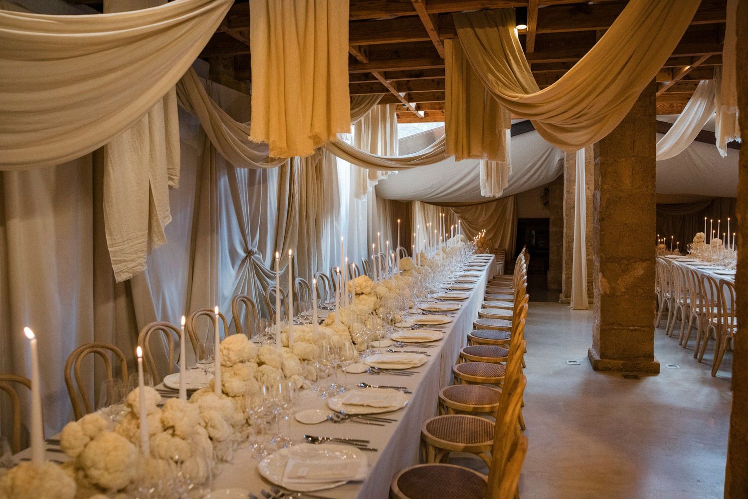 Long banquet table with white draping and floral decor in an elegant rustic venue.