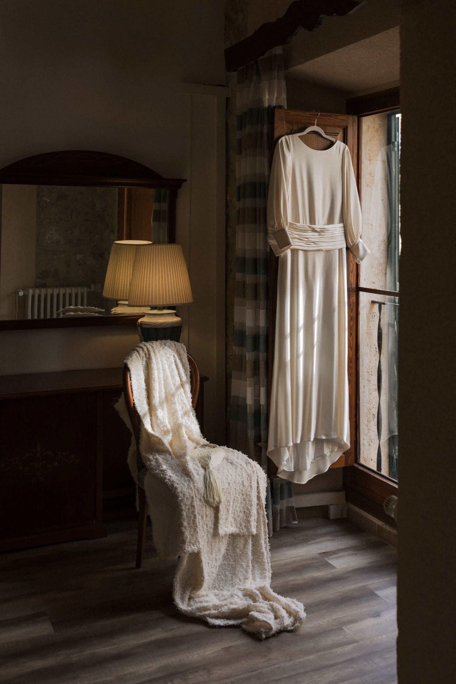 White wedding dress and veil hanging in dimly lit room with vintage furniture.