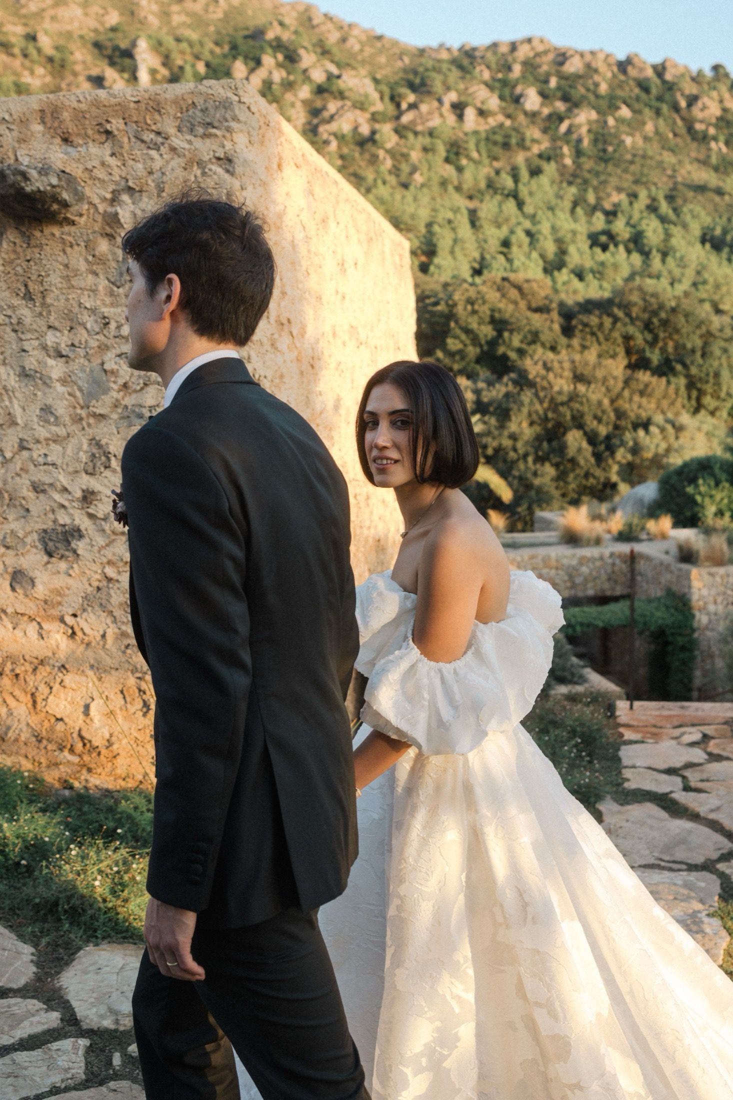 A couple in wedding attire poses against a stone wall with mountains in the background during golden hour.