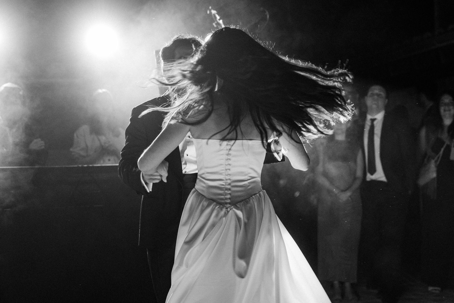 A person in a white dress dances energetically with hair flying at a dimly lit event.