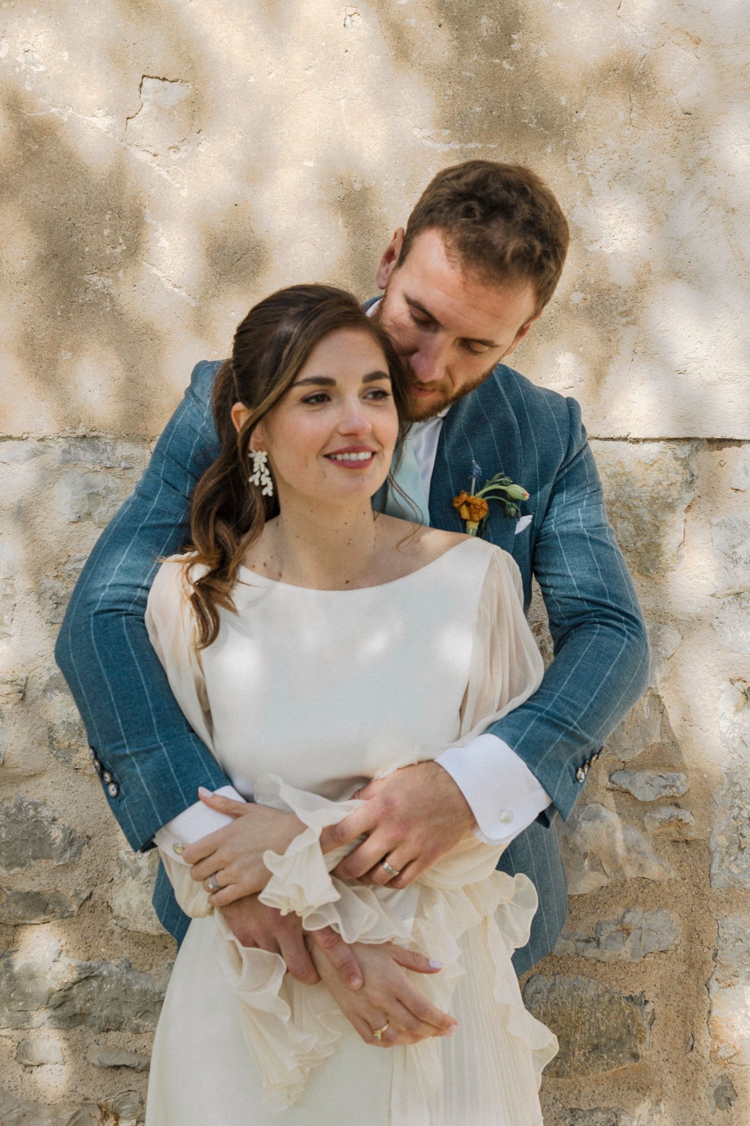 A romantic couple in wedding attire embraces against a textured stone wall with dappled sunlight.