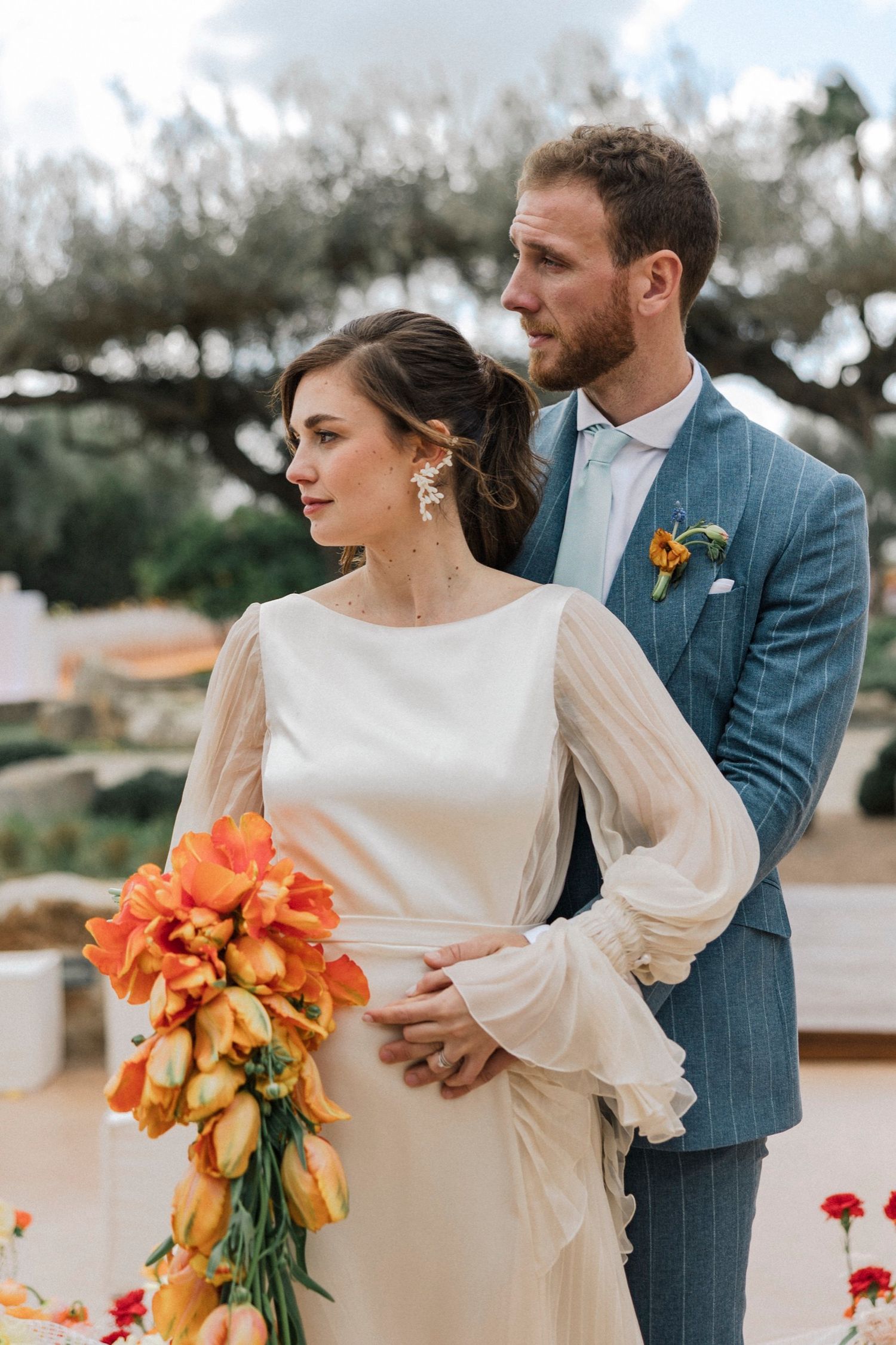 A wedding couple in modern attire with the bride holding vibrant orange lilies against a bright outdoor backdrop.