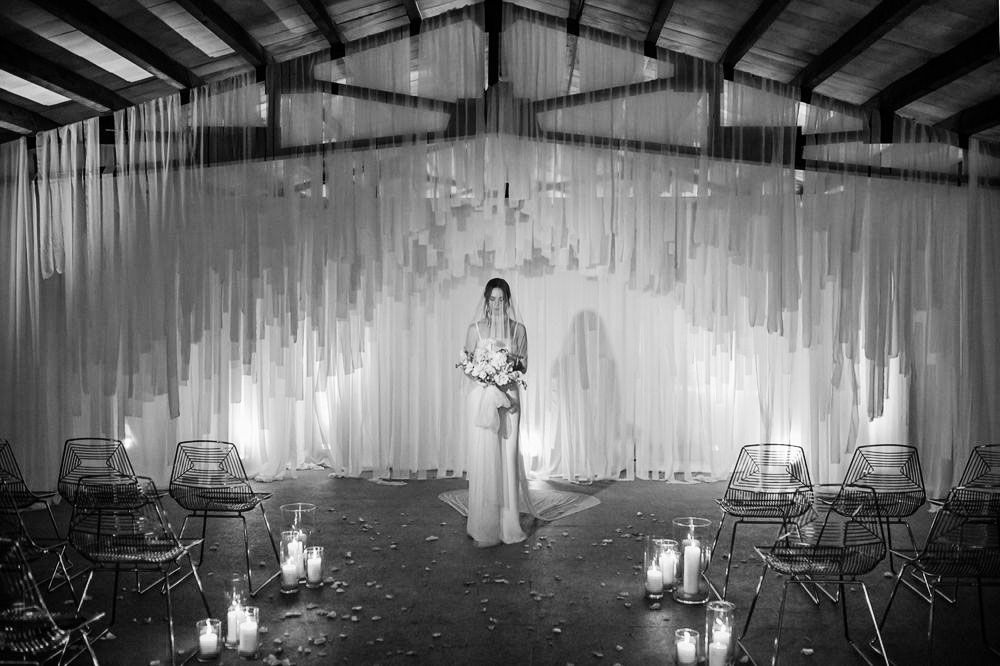 A dramatic black and white wedding scene with white draping, lanterns, and metal chairs in an indoor venue.