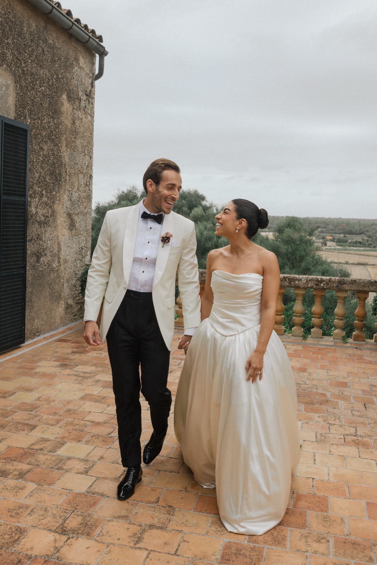 A couple in wedding attire share a joyful moment on a terracotta balcony overlooking a countryside landscape.