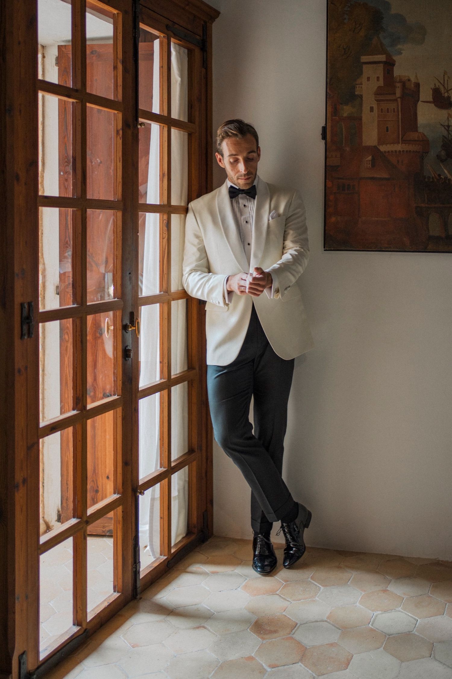 Person in a white dinner jacket and dark pants standing by large windows in an elegant interior space.
