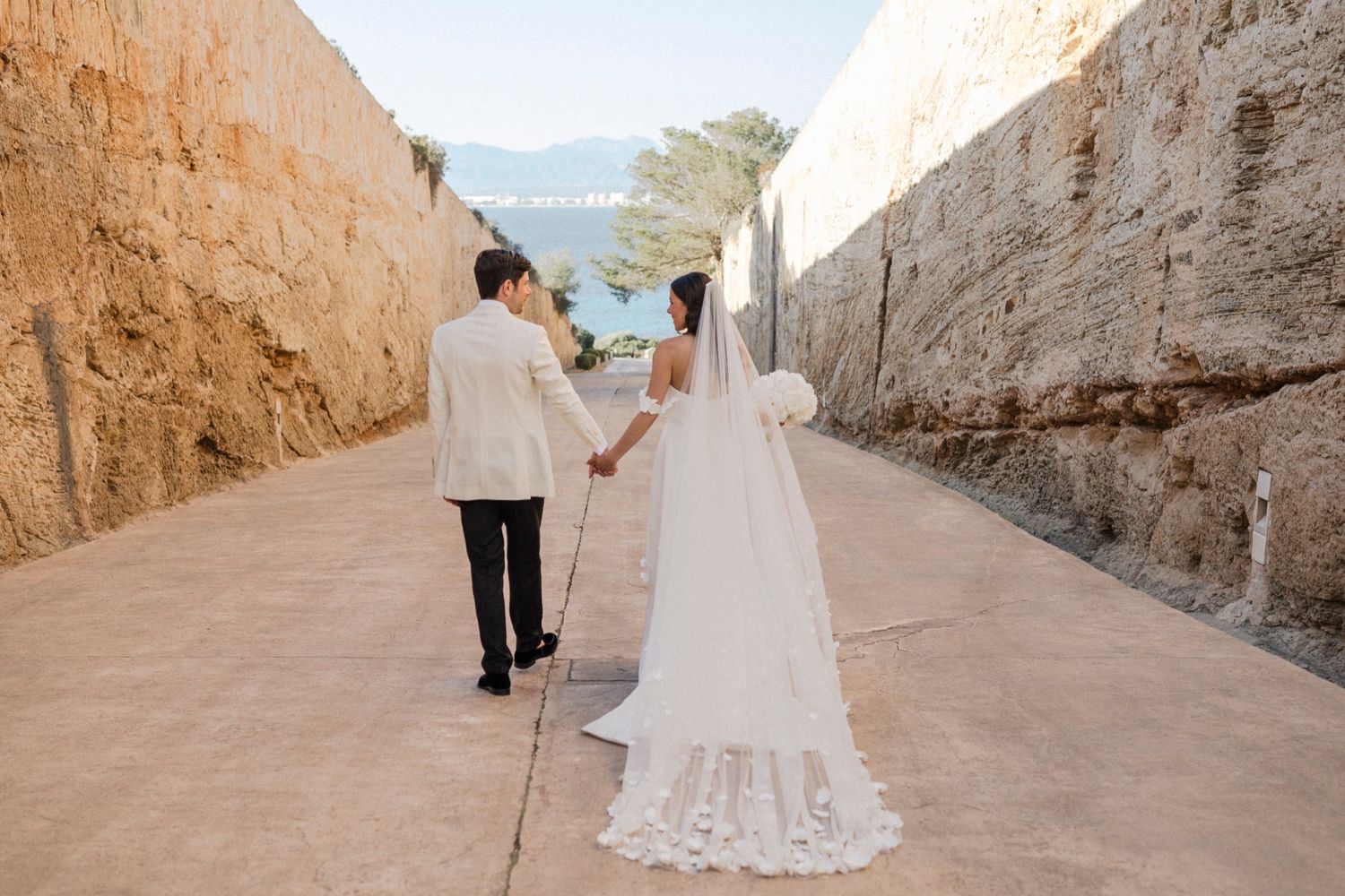Bride and groom holding hands walking through stone corridor in wedding attire, cathedral veil trailing behind.