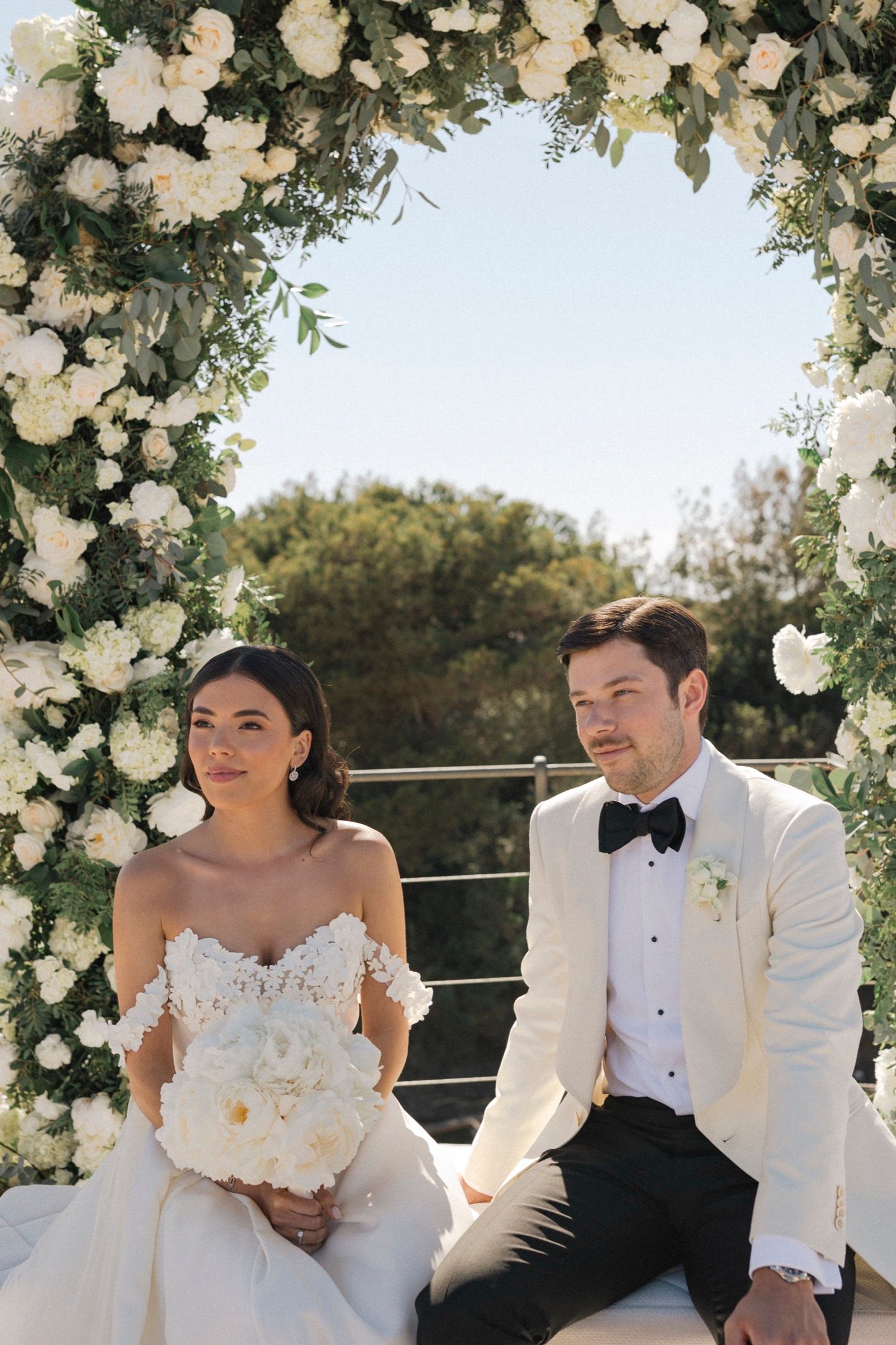 Bride and groom sitting under a white floral arch outdoors, bride holding a white bouquet, groom in white tuxedo jacket.