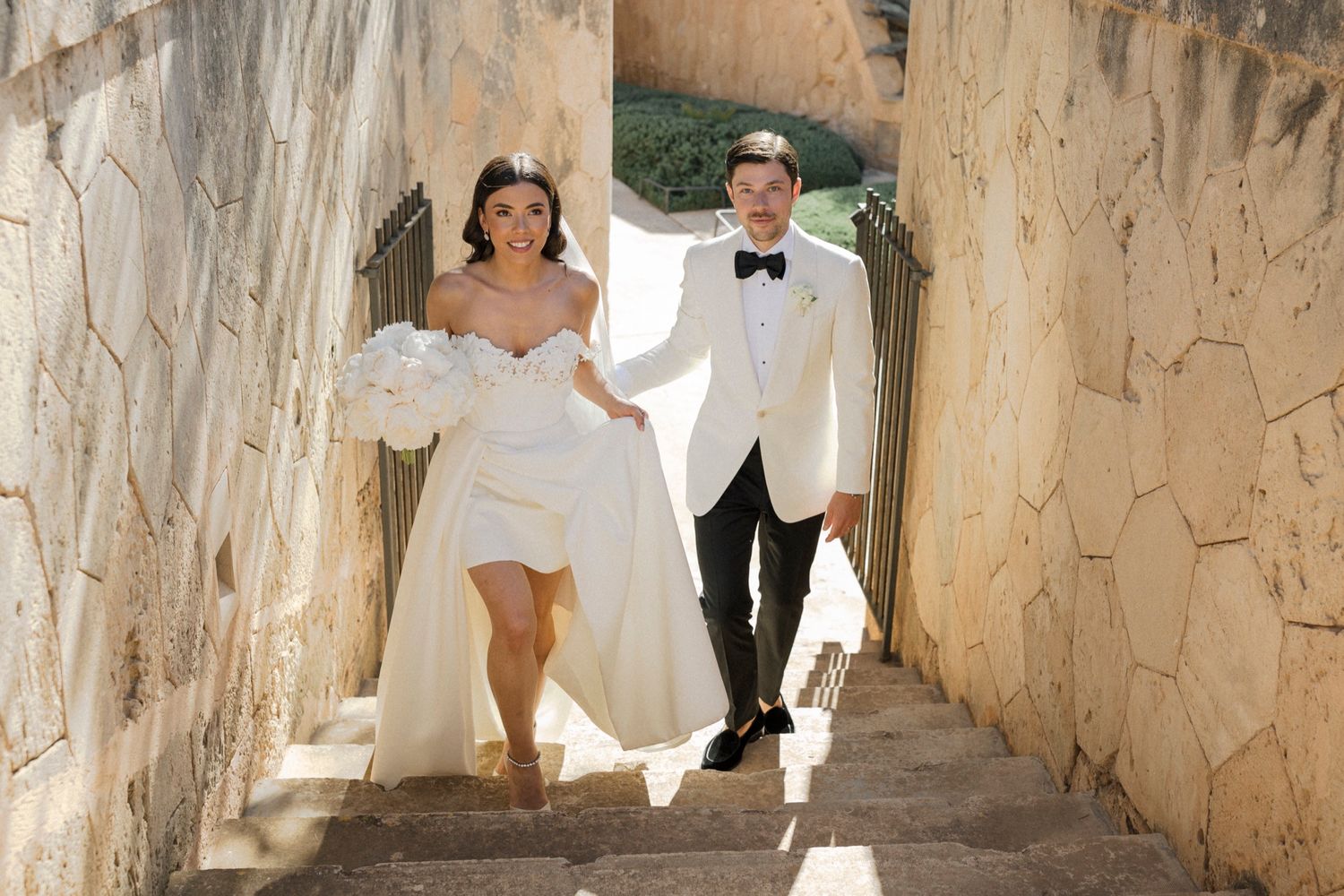 Bride in white off-shoulder gown and groom in white tuxedo walking down stone steps holding hands.