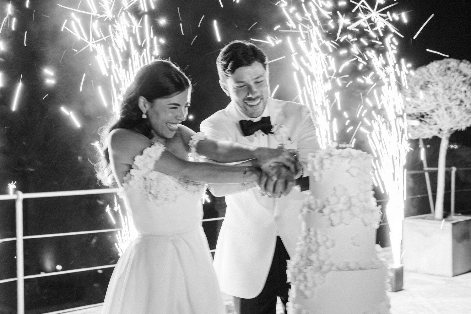 Bride and groom cutting wedding cake with sparklers in black and white photo.