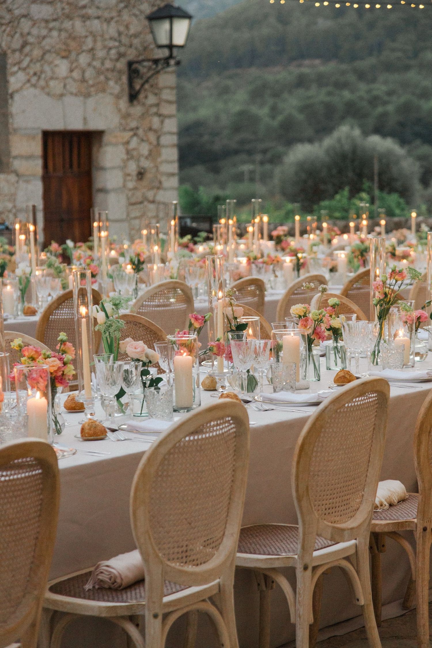 Elegant outdoor wedding reception table with rattan chairs, taper candles and floral centerpieces against stone wall backdrop.