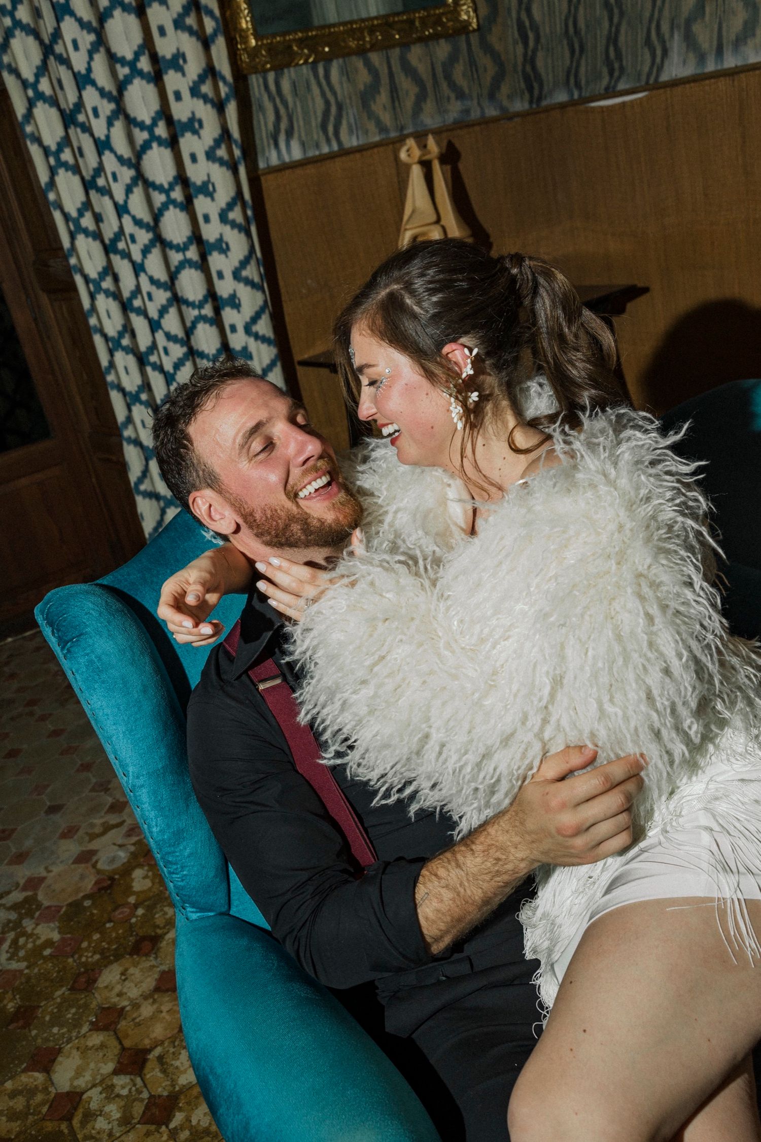 A couple shares a joyful intimate moment on a teal chair, with the woman wearing a white feathered outfit.