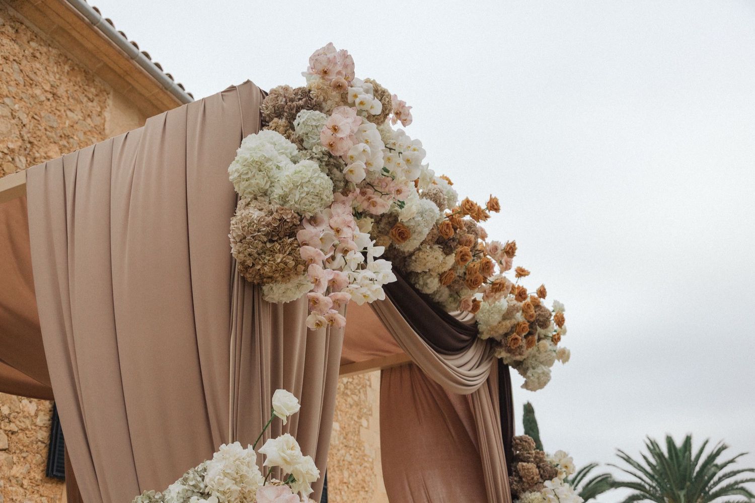 Elegant outdoor wedding arch decorated with blush and cream flowers and flowing taupe fabric against a cloudy sky.