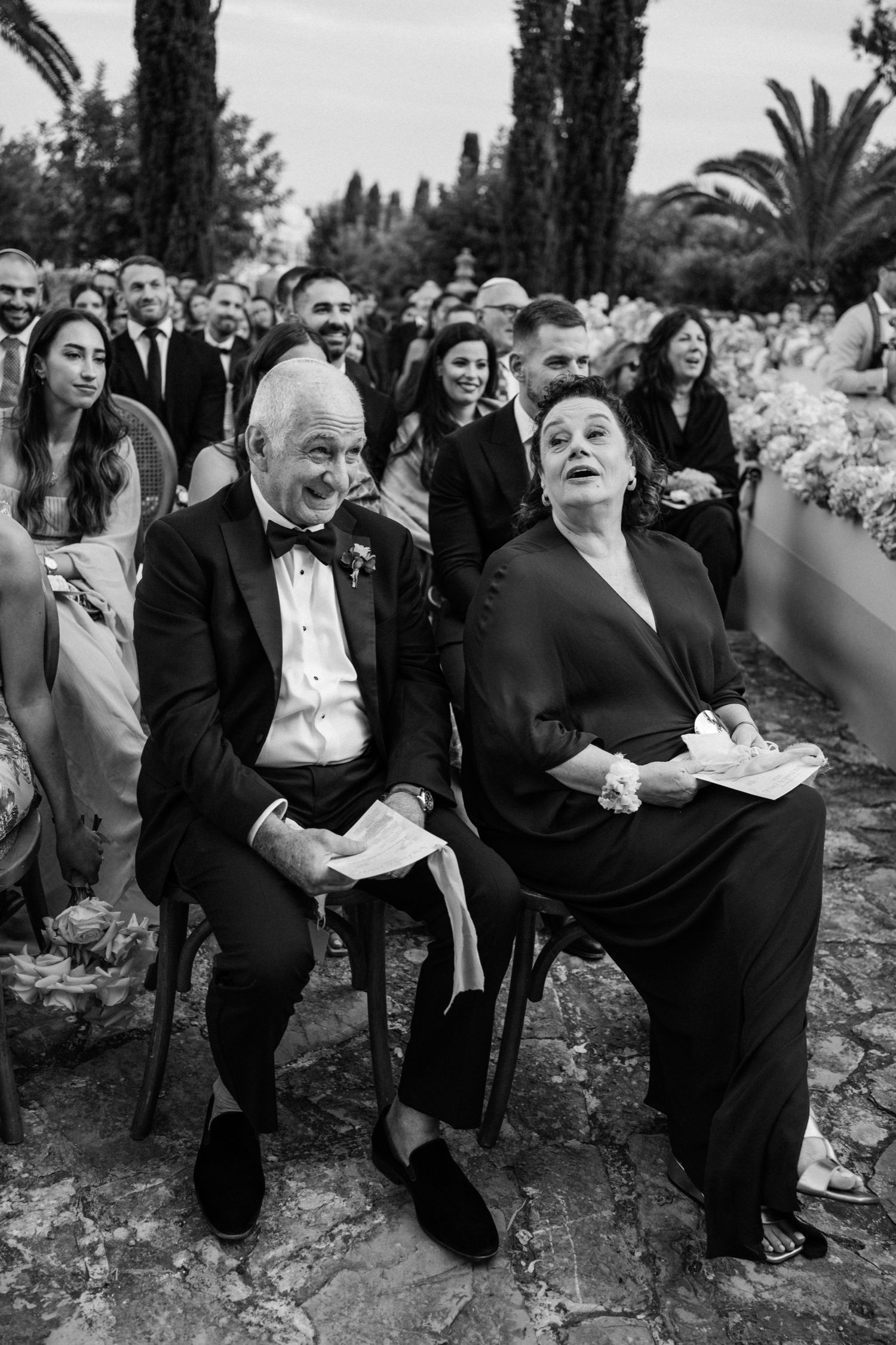 Wedding guests seated outdoors among cypress trees and palm trees watch an emotional ceremony in black and white.