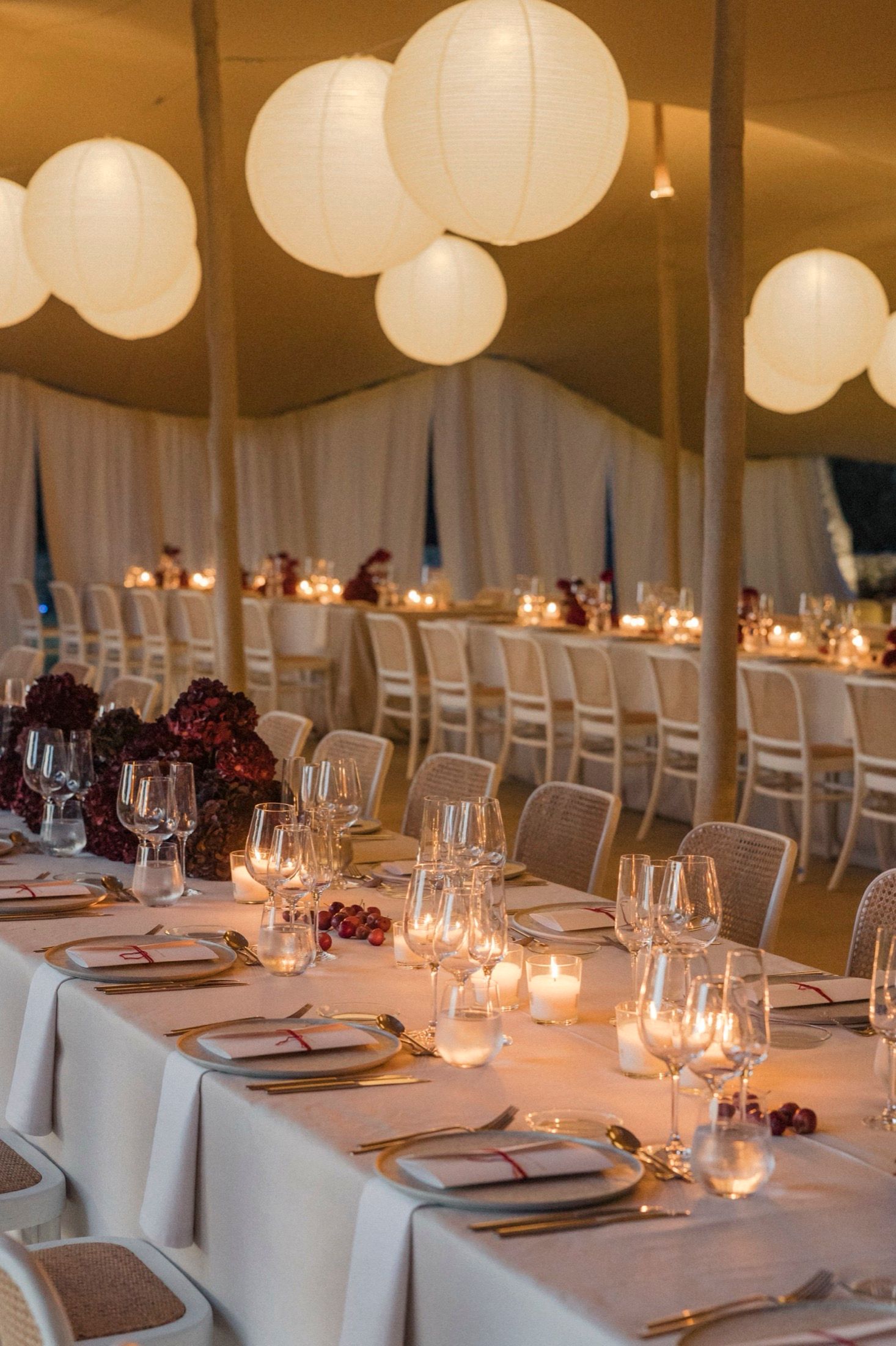 Elegant wedding reception table setting with white linens, candlelight, wine glasses and deep burgundy floral centerpieces.