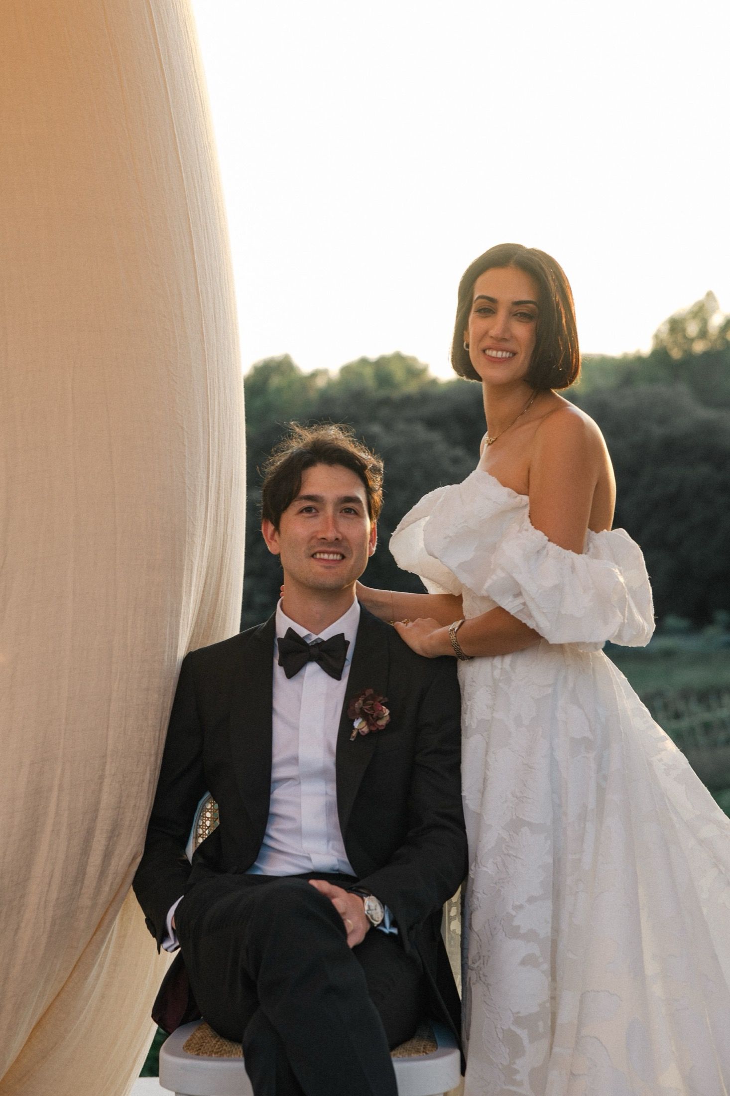 A newlywed couple laughs together while seated between elegant white drapes at their wedding venue.