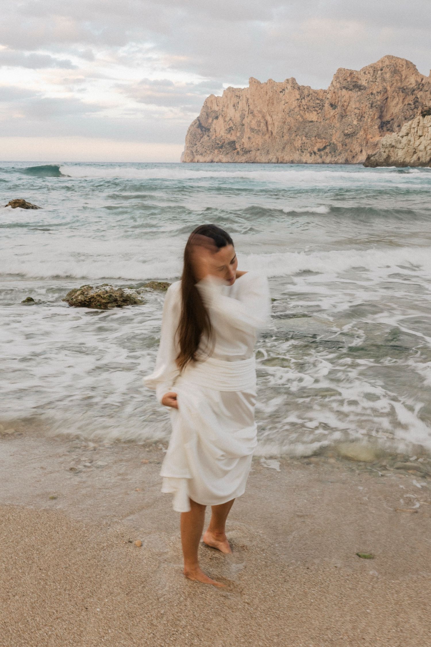 Woman in white dress twirls on beach with waves and coastal cliffs in background.
