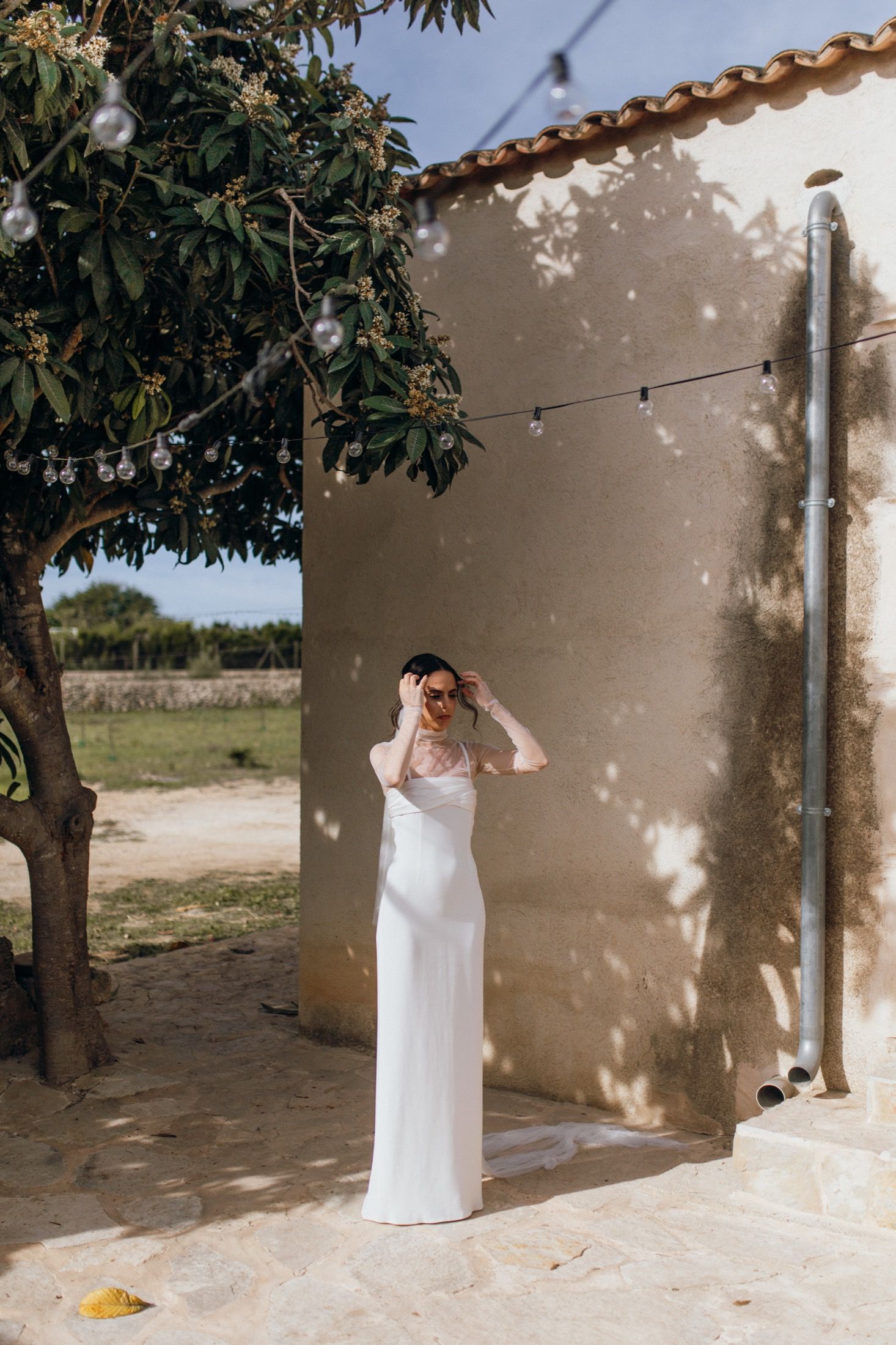 A white wedding dress against a rustic beige wall with string lights and a tree casting dappled shadows in natural sunlight.
