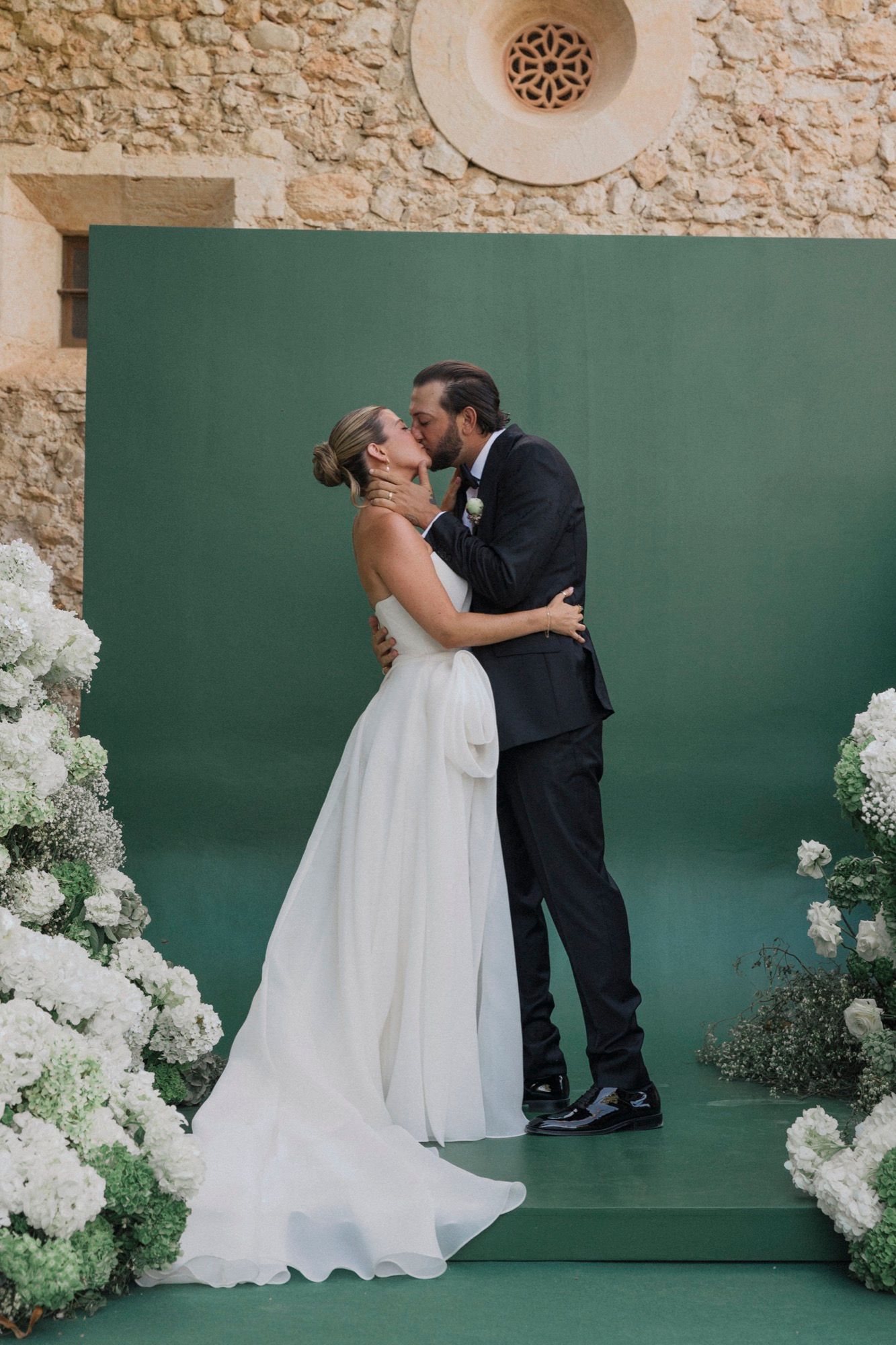 Romantic wedding kiss on green platform surrounded by white hydrangea arrangements.