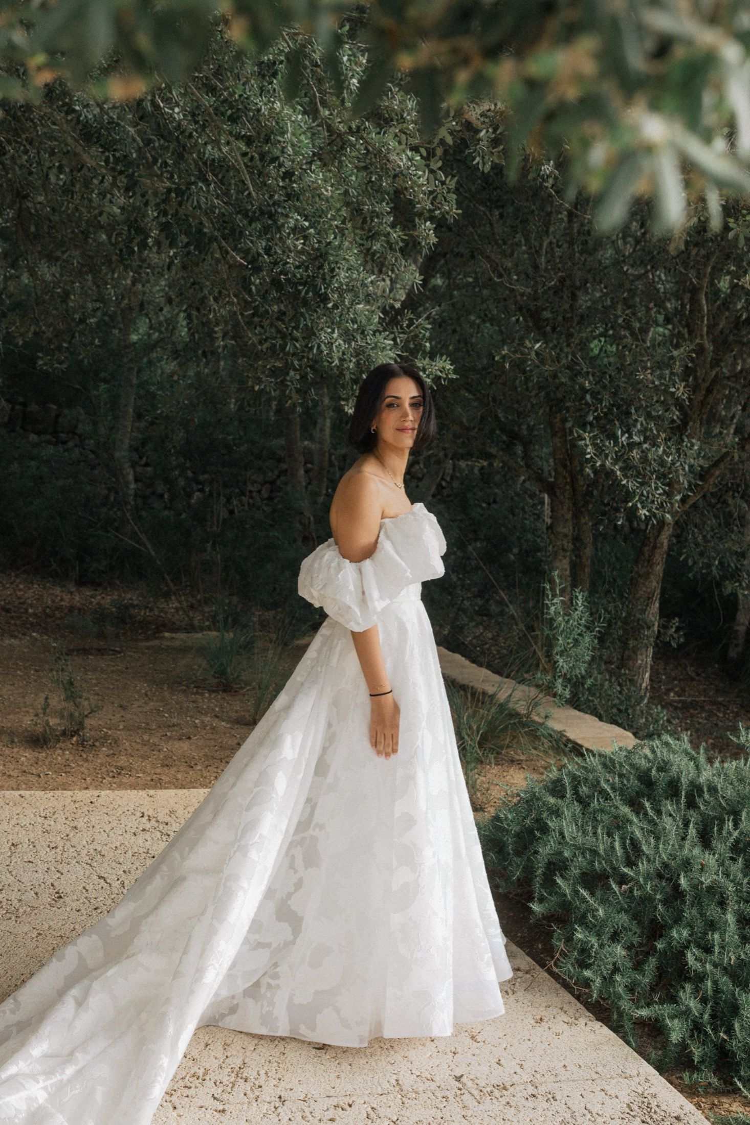 A flowing white wedding dress with puff sleeves and lace details photographed in a garden setting.