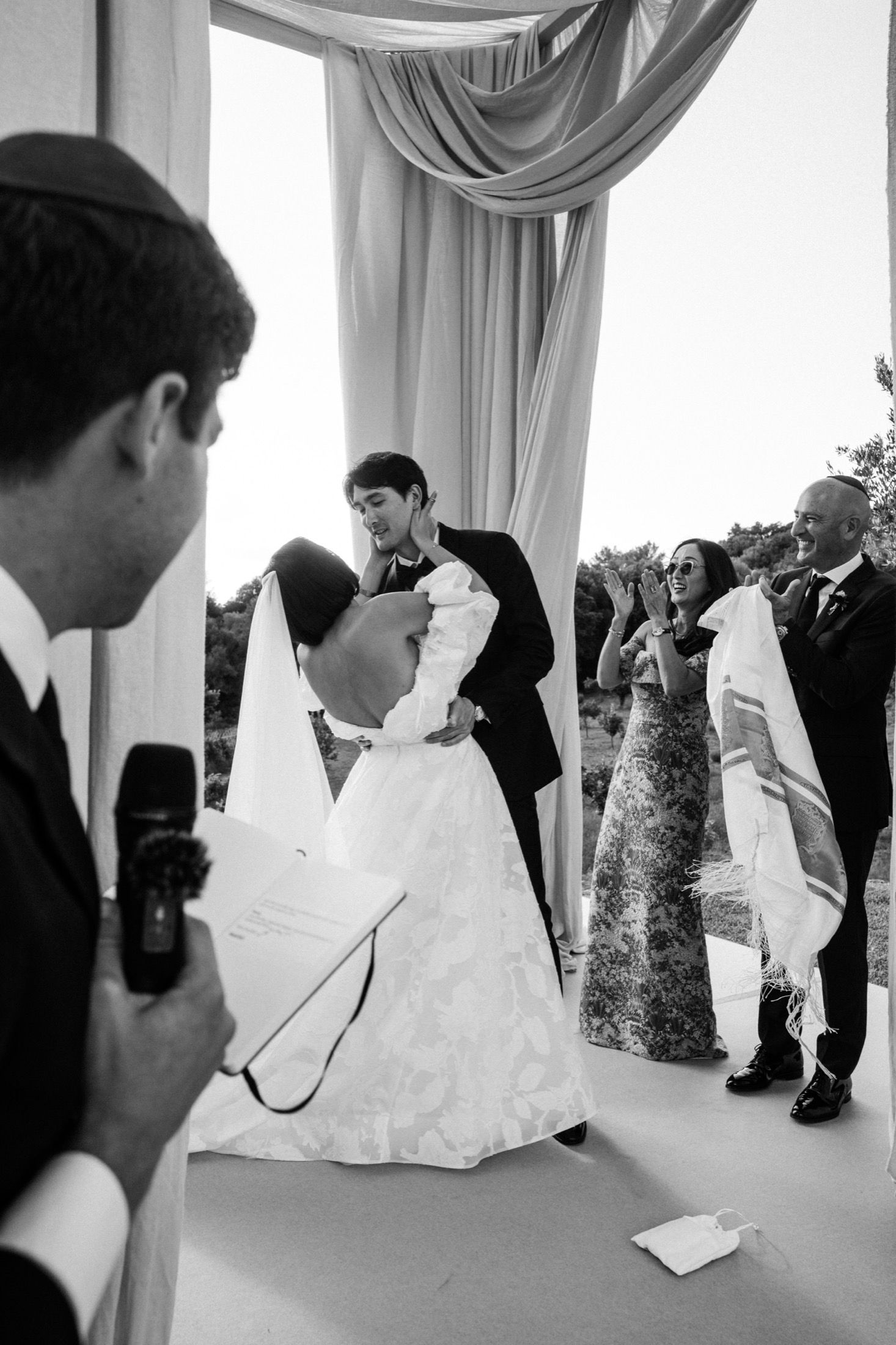 Black and white wedding ceremony photo showing couple embracing under draped gazebo while guest speaks into microphone.