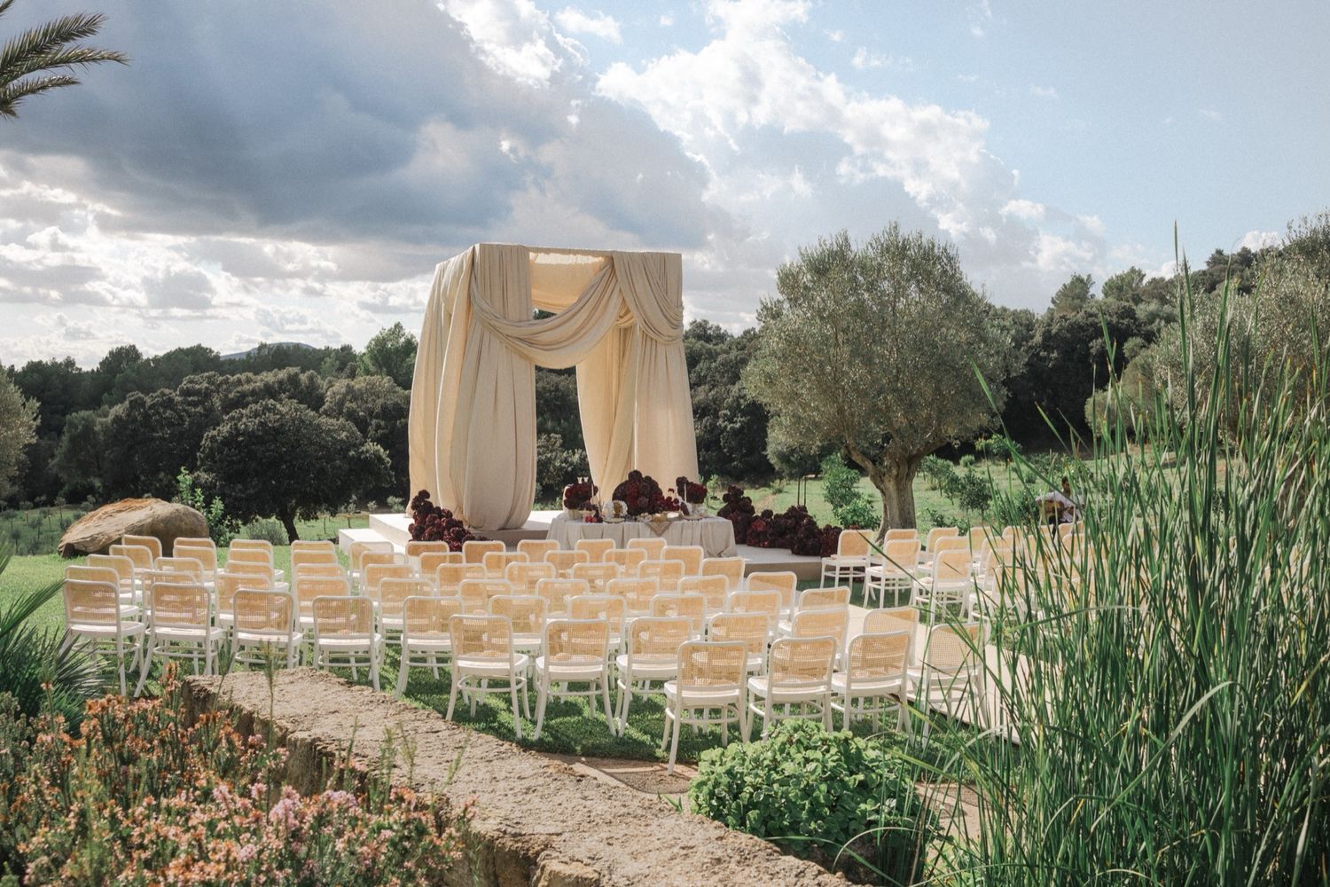 Outdoor wedding ceremony setup with draped fabric arch and white chairs on gravel path surrounded by lush gardens.