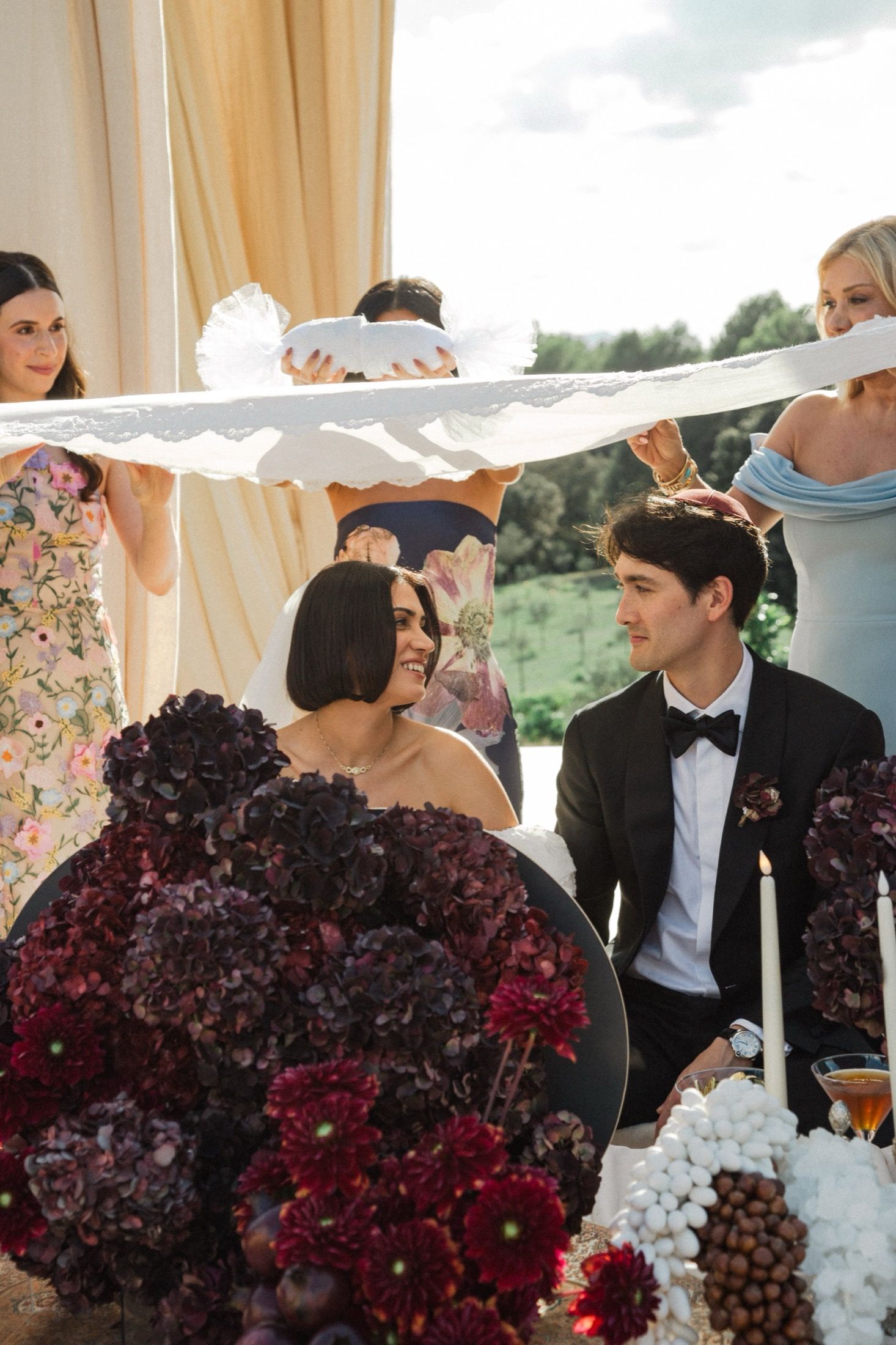 Wedding reception table decorated with dramatic burgundy and white floral arrangements under an elegant white tent.