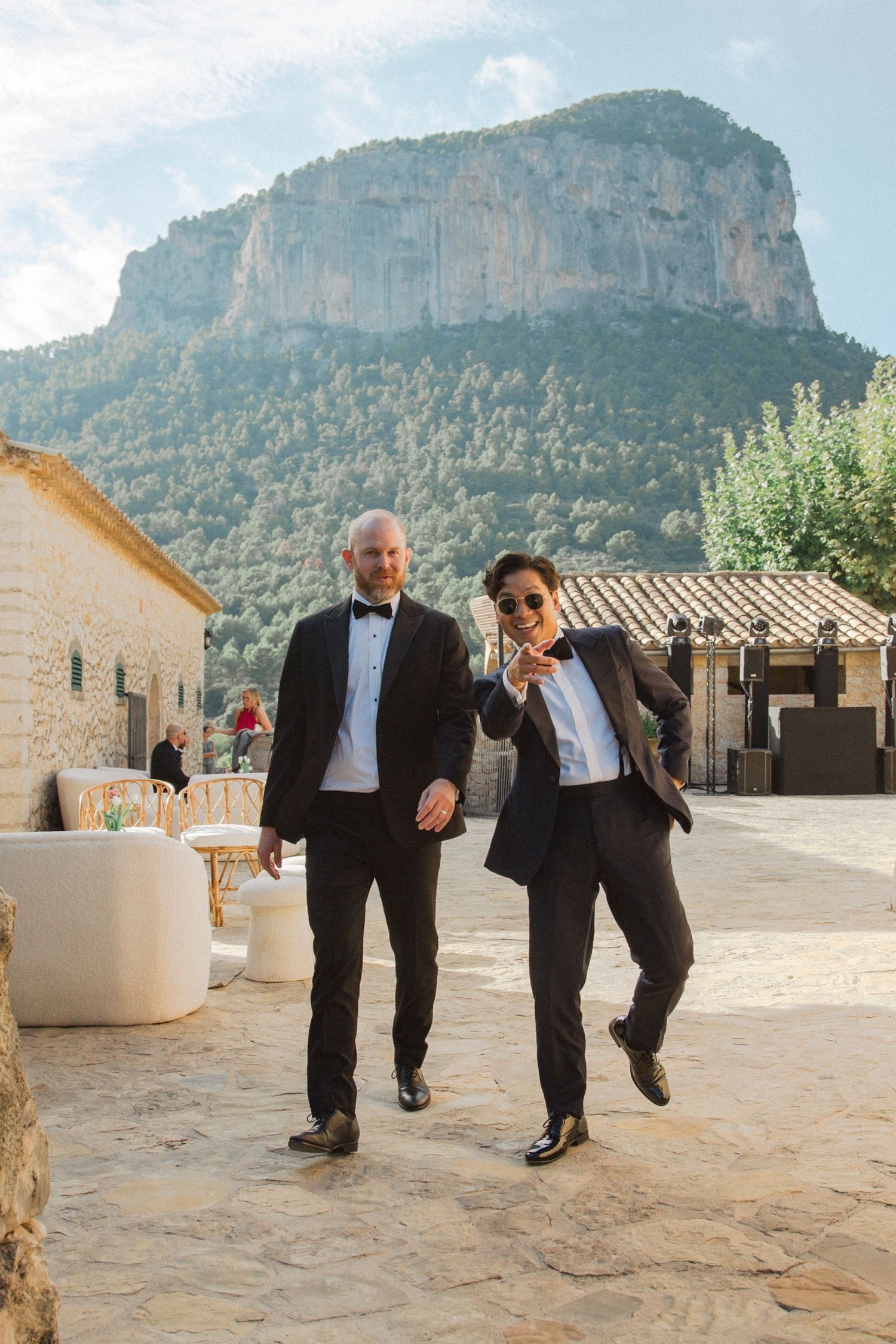 Two people in formal attire perform a playful dance sequence against a backdrop of rustic buildings and mountains.