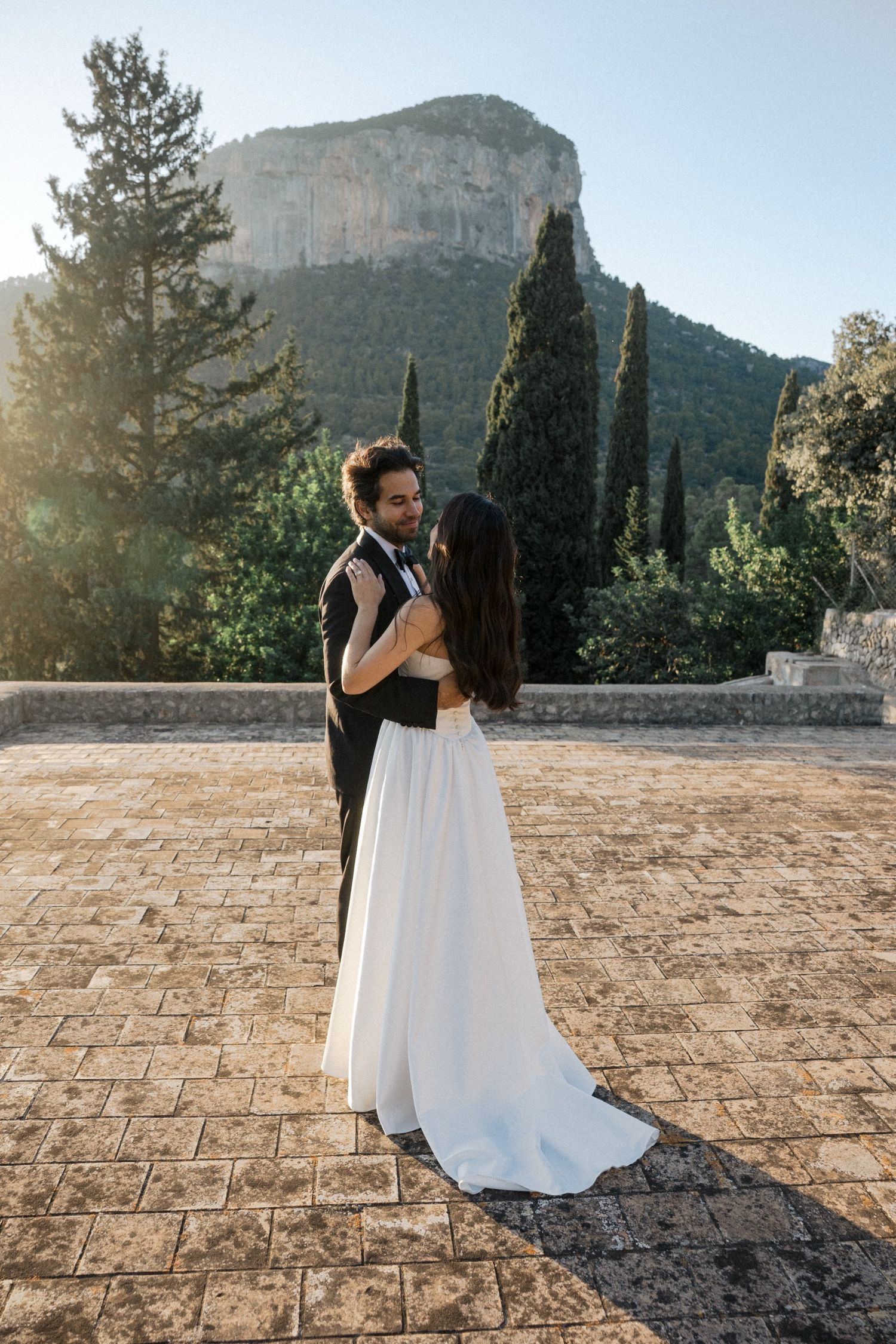 A couple shares a romantic embrace at sunset on a stone patio with mountains and cypress trees in the background.
